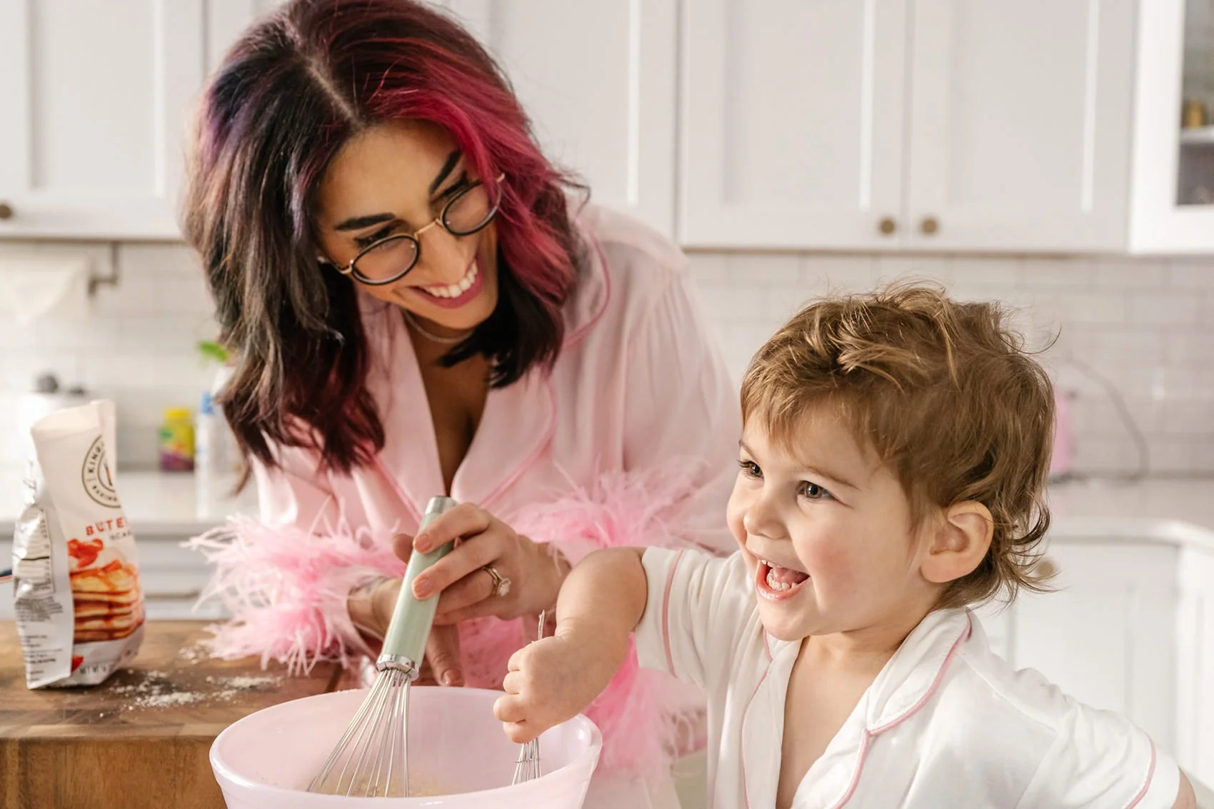 Happy mother with pink hair and pink pajamas with feather trim, smiling while helping young daughter mix ingredients in bright Maplewood NJ kitchen.