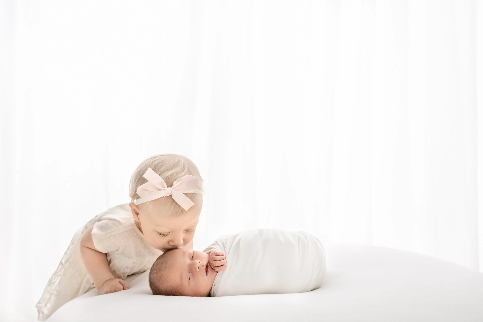 Toddler sister wearing cream dress and bow in her hair gently kisses her swaddled newborn baby brother