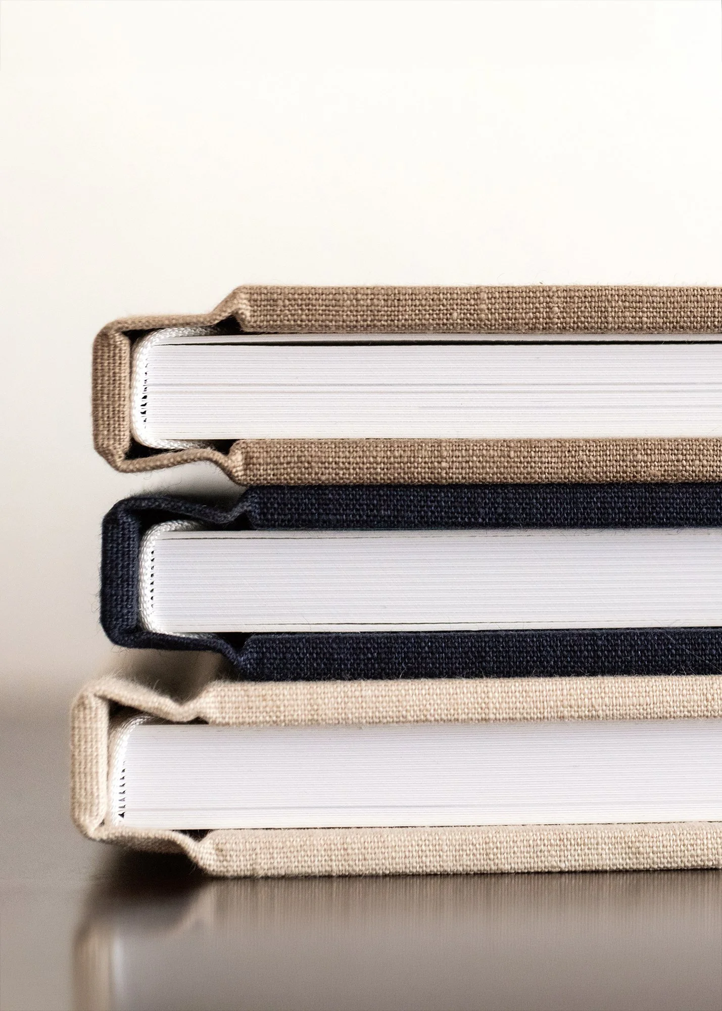 Close-up view of stacked hardcover photo albums with fabric covers in beige, navy blue, and light brown, placed on a reflective surface.