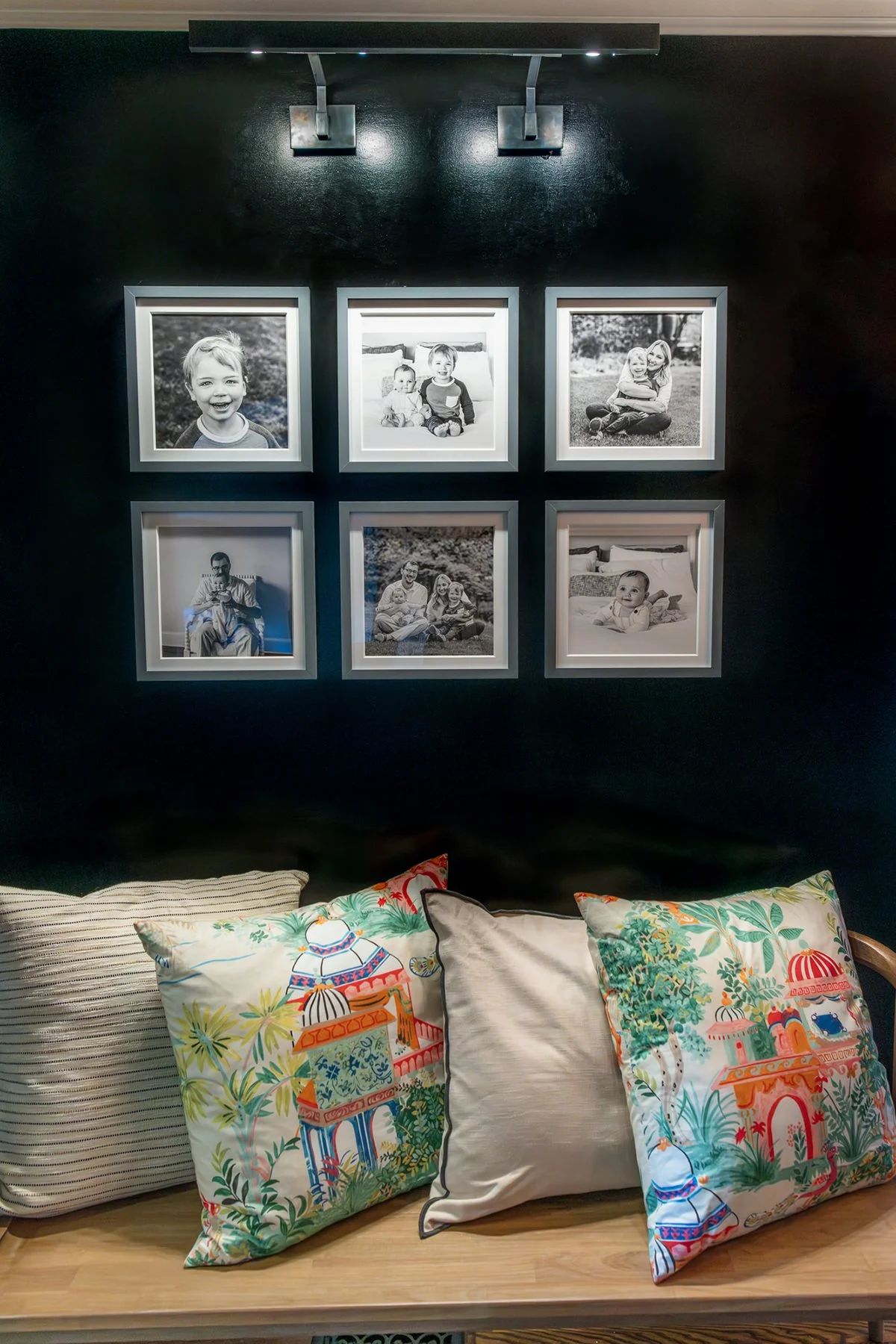 Modern black and white framed family photo gallery on black wall in South Orange, with overhead lights and bench below.