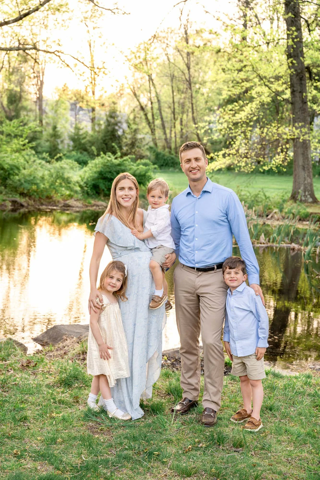 A family of six standing outdoors by a pond with green trees in the background, smiling at the camera during sunset.