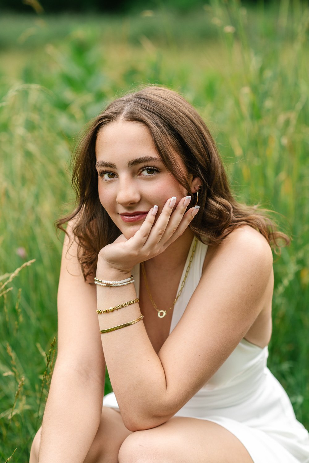 Natural high school senior portrait with brunette wearing a white dress and gold jewelry, crouched, with her face gently resting on hand.