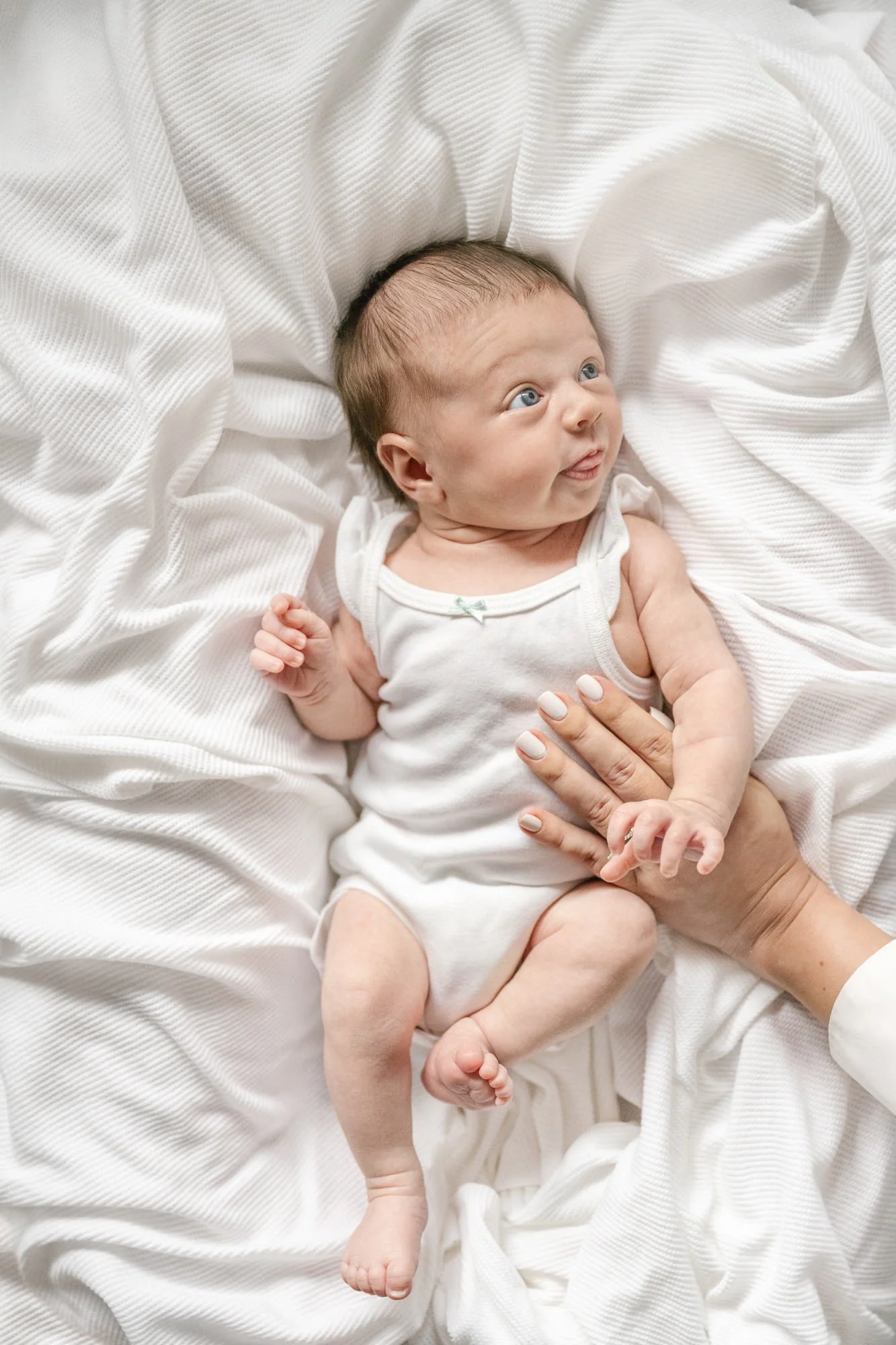 Darling newborn sticks out her tongue at mom off camera. Baby dressed in classic white lays on a timeless colored blanket. Wide awake blue-eyed baby girl. #centralNewJerseyphotographer #inhomeportraits #newbornphotoshoot #NewJerseyPhotographers