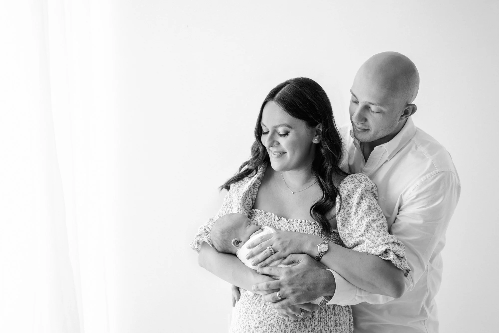 Captivating black and white moment: A newborn cradled in their mother's arms, with the father standing protectively behind, radiating a sense of love and strength in a Nicole Hawkins photography studio session in New Jersey. #NicoleHawkinsphotography