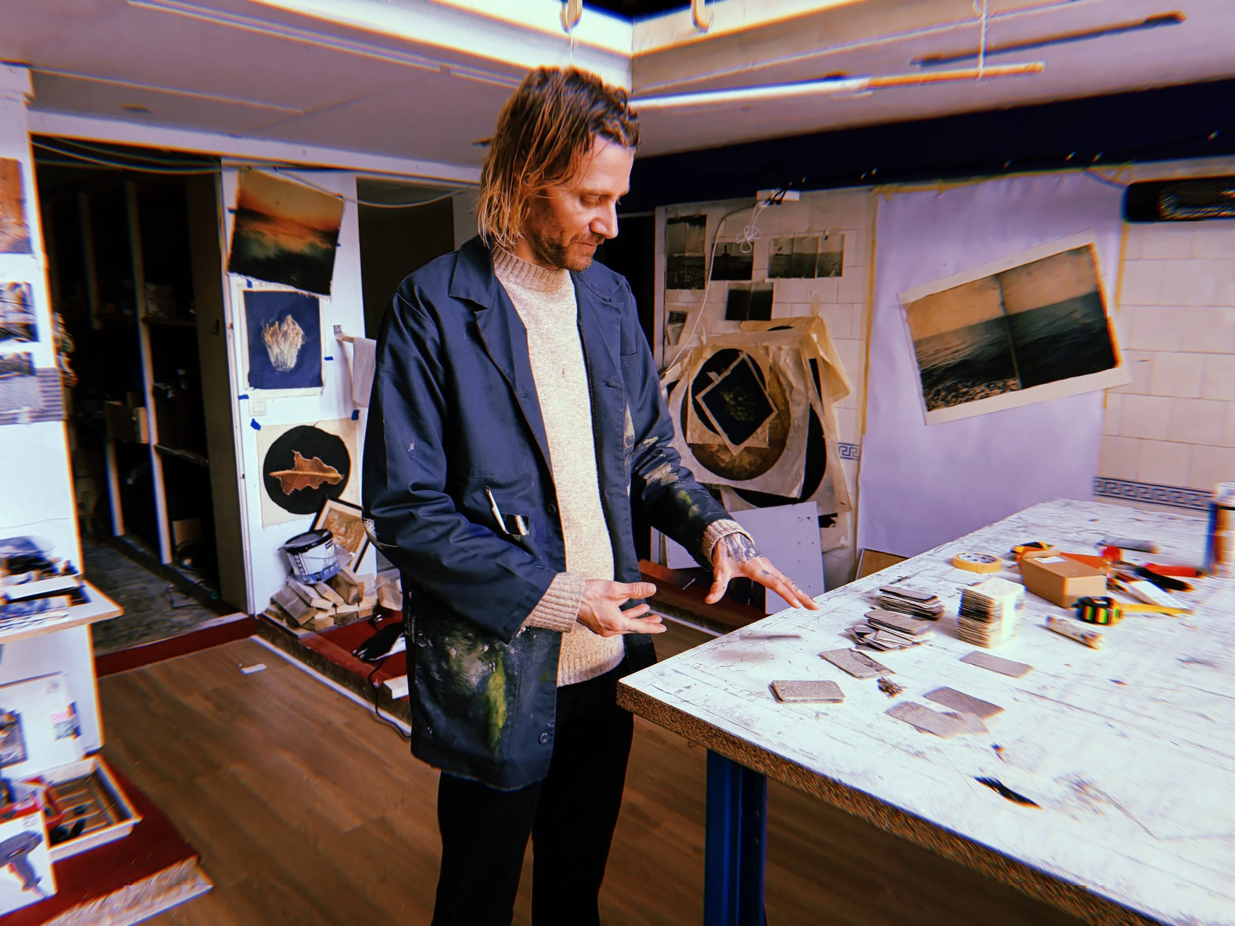 An artist standing at a studio worktable with loose photographic cards and stacks of fragments.