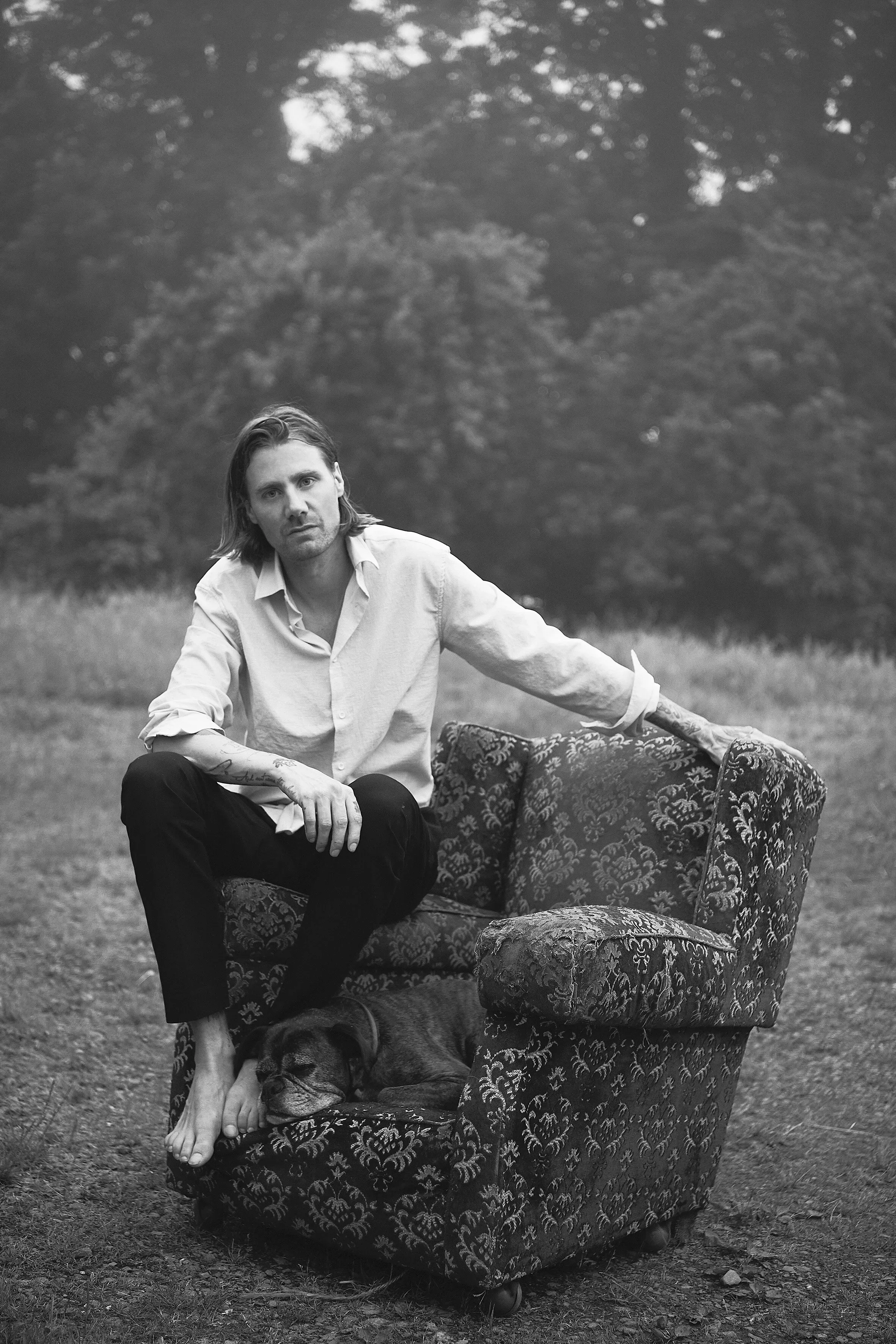 Black-and-white portrait of Joost Vandebrug seated outdoors on an upholstered chair, with a dog resting at his feet.