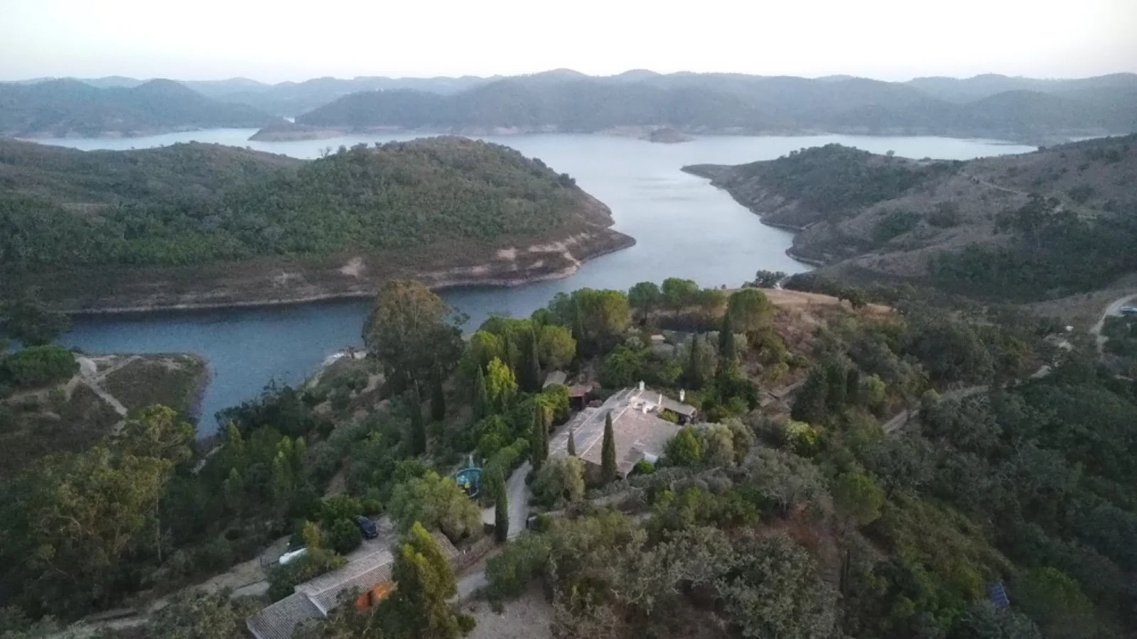 Aerial view of a landscape with a large body of water surrounded by hilly terrain, greenery, and a few buildings and trees in the foreground.