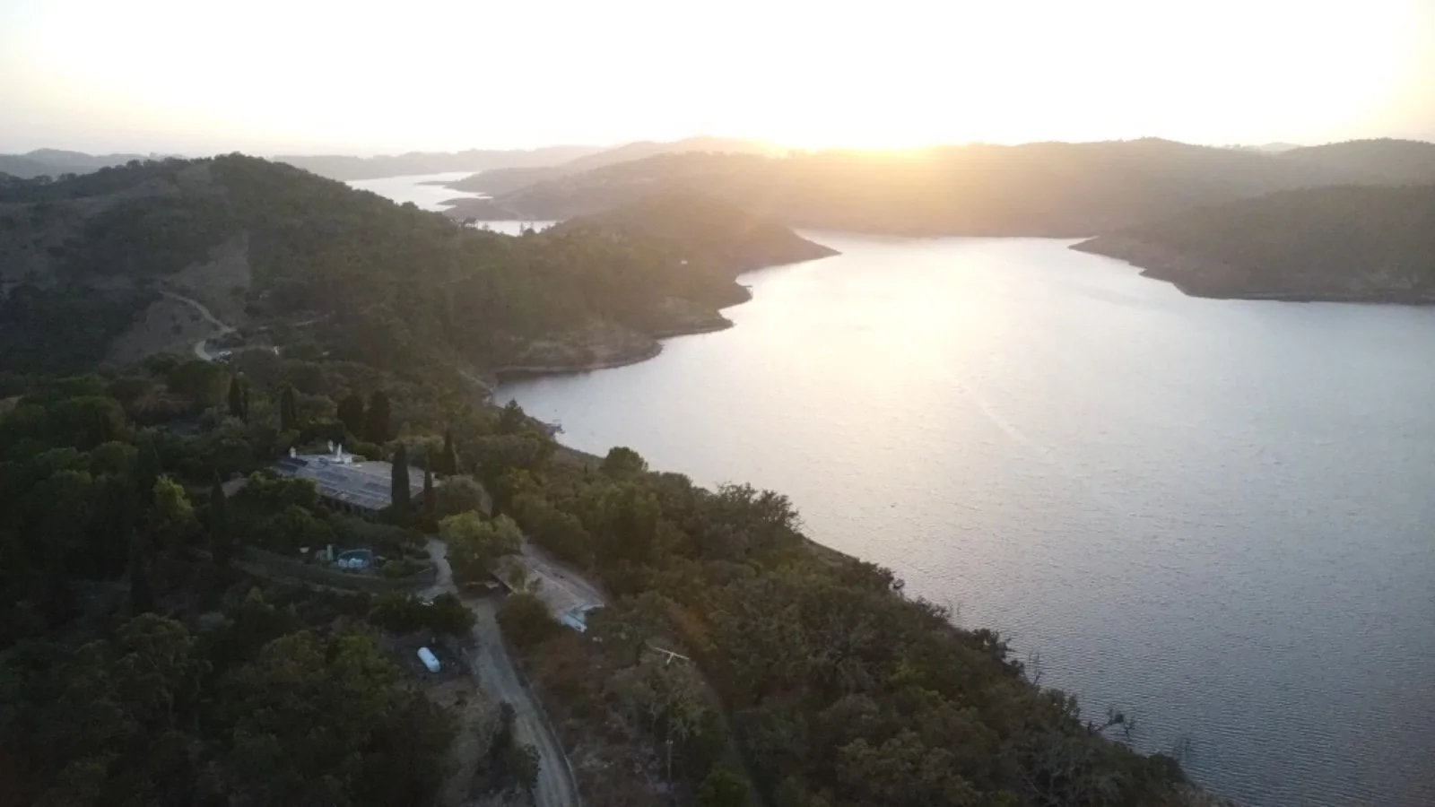 Aerial view of a large lake surrounded by green hills during sunset with a small road and buildings in the foreground.
