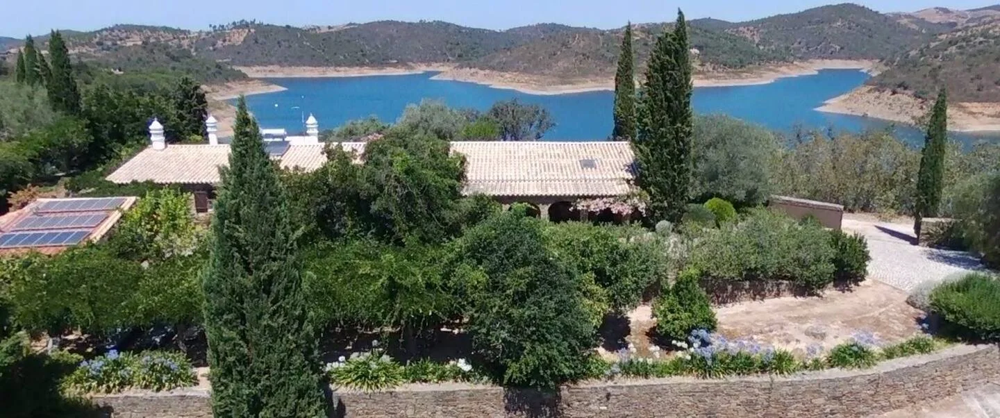 A house surrounded by trees and shrubs with a lake and rolling hills in the background.