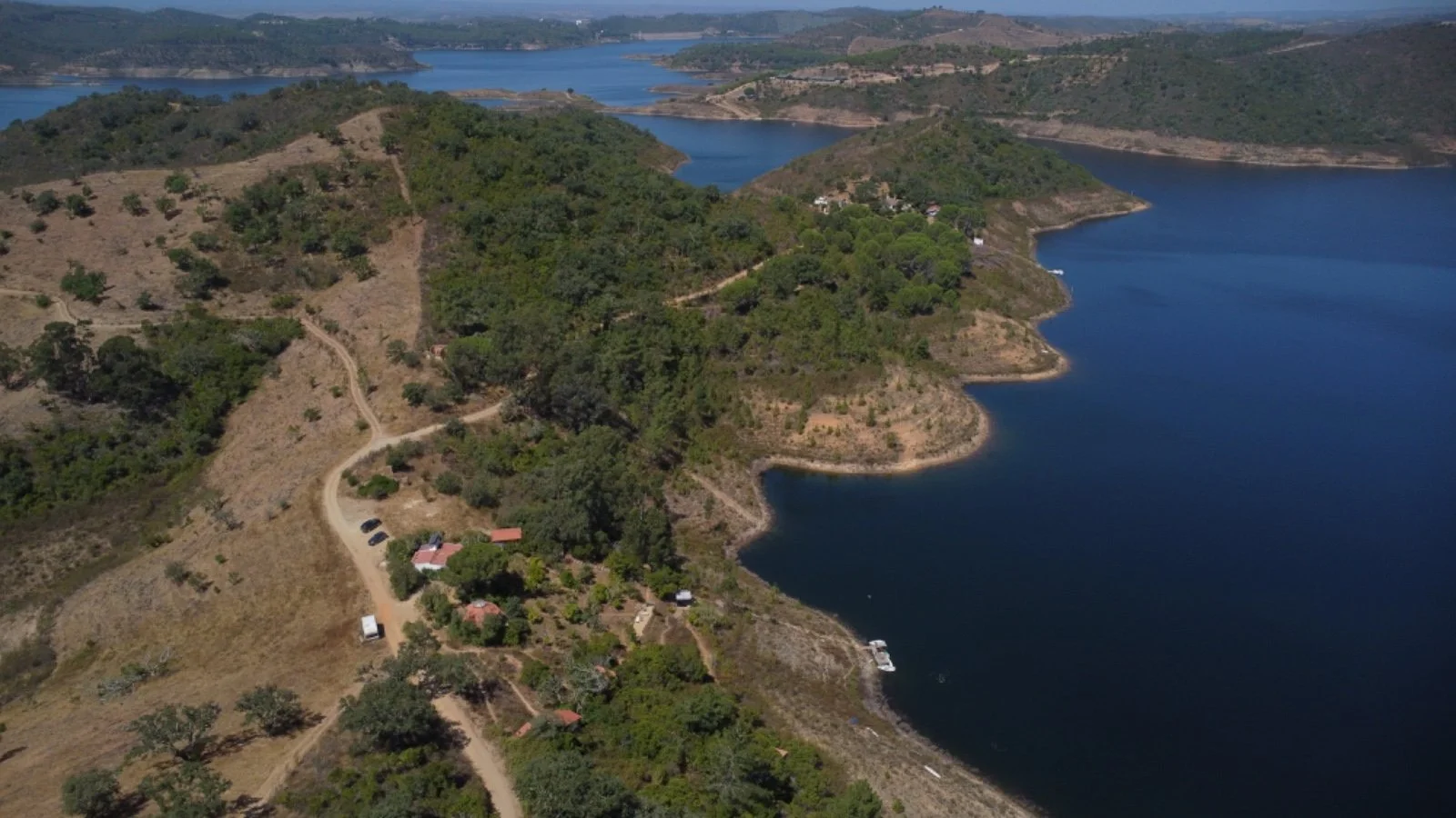 Aerial view of a hilly landscape with a large body of water, with several small houses and a winding road along the shoreline surrounded by trees and dry terrain.