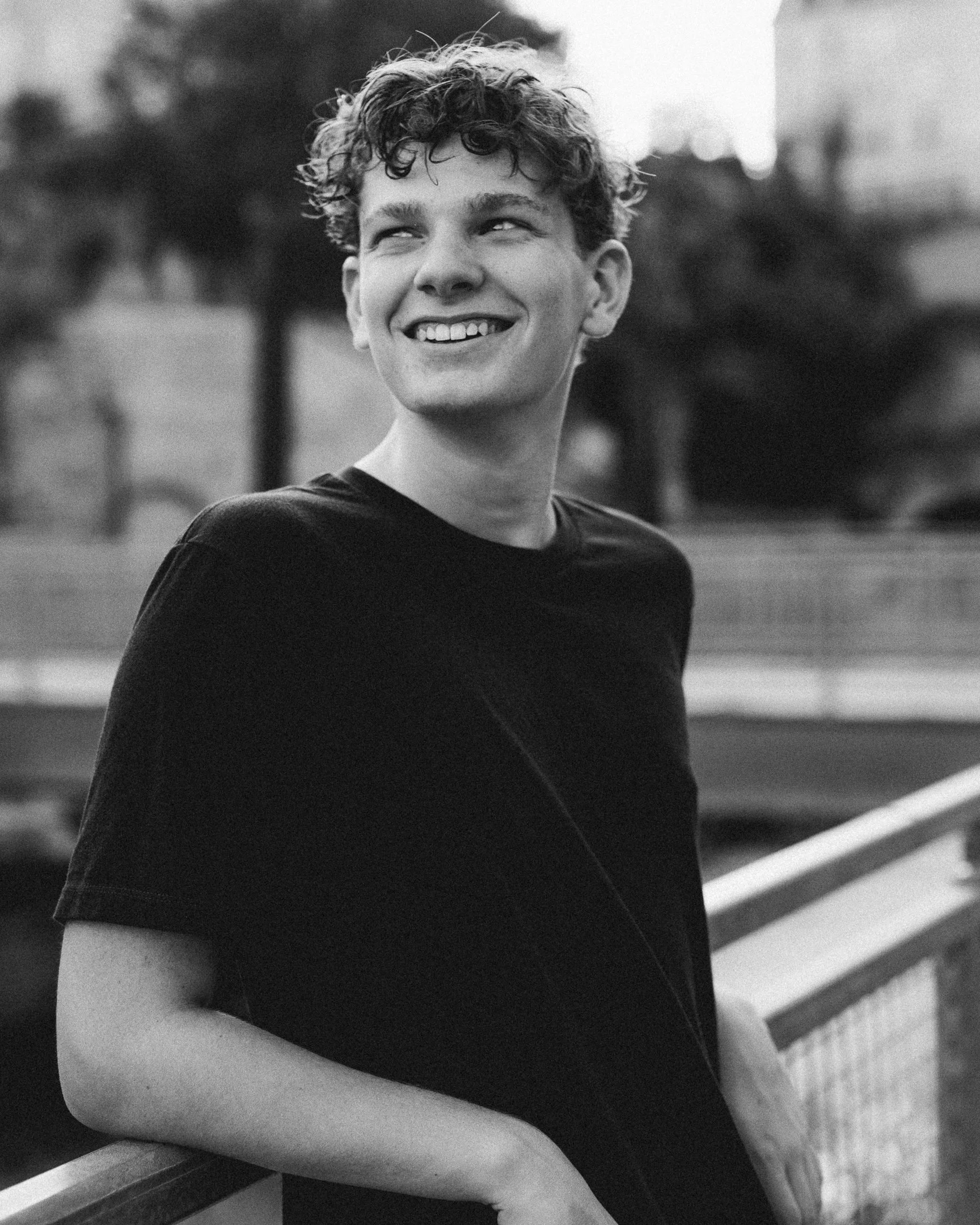 A young man with curly hair smiling and looking towards the left, standing outdoors in front of a railing, in black and white.