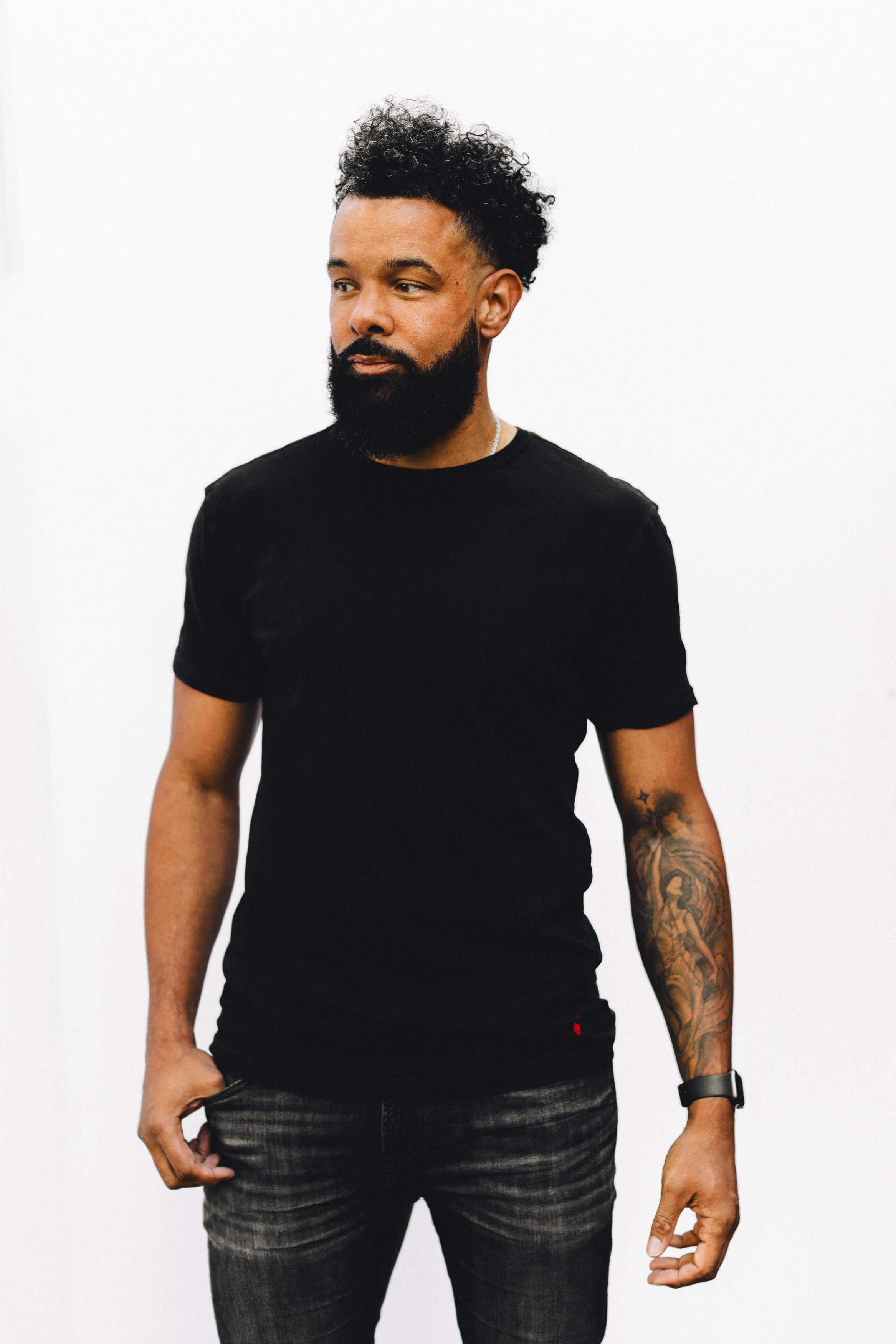A man with dark curly hair and a beard wearing a black T-shirt and jeans standing against a plain white background.