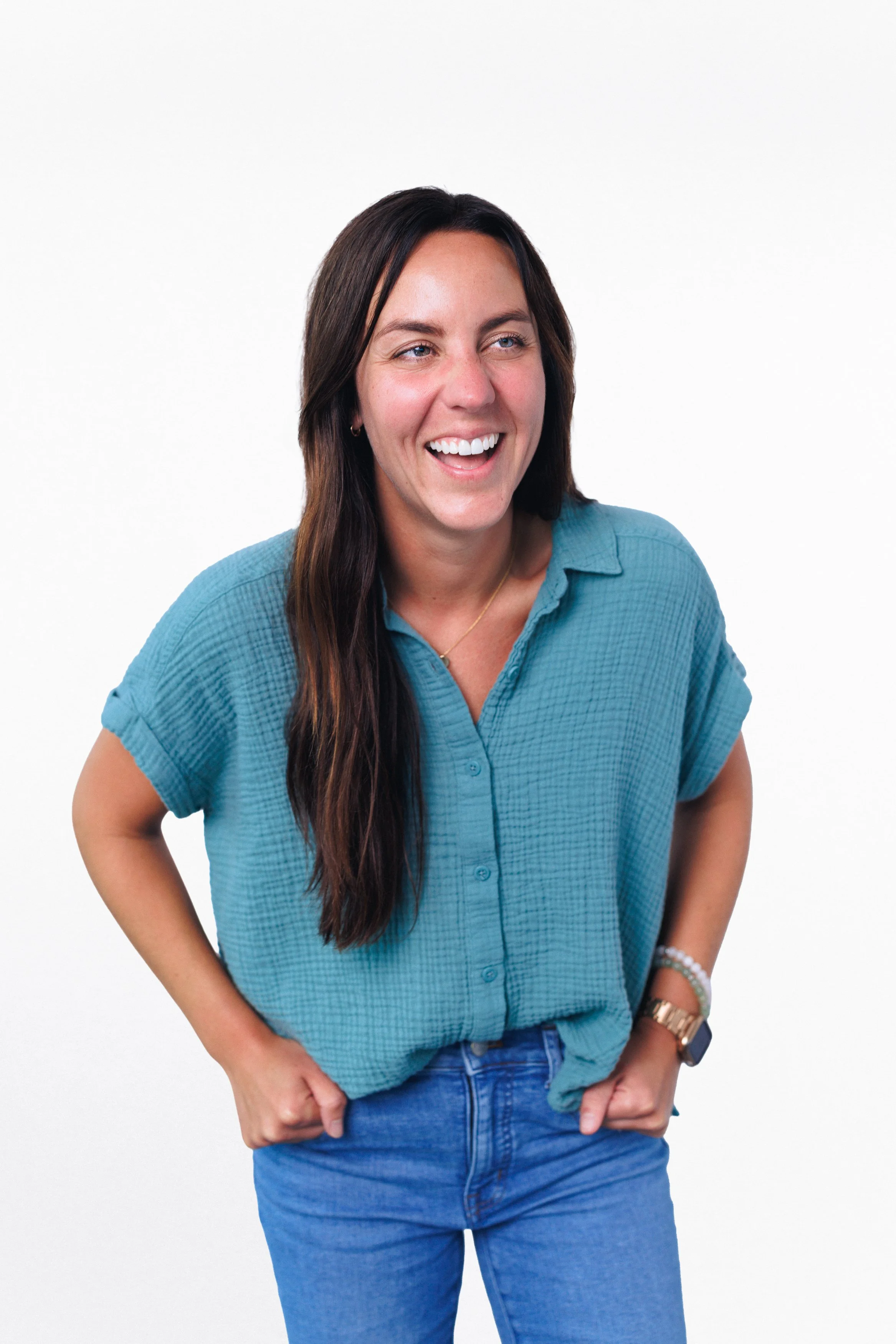 A woman with long brown hair, wearing a turquoise short-sleeve button-up shirt and blue jeans, smiling and looking to her left against a plain white background.