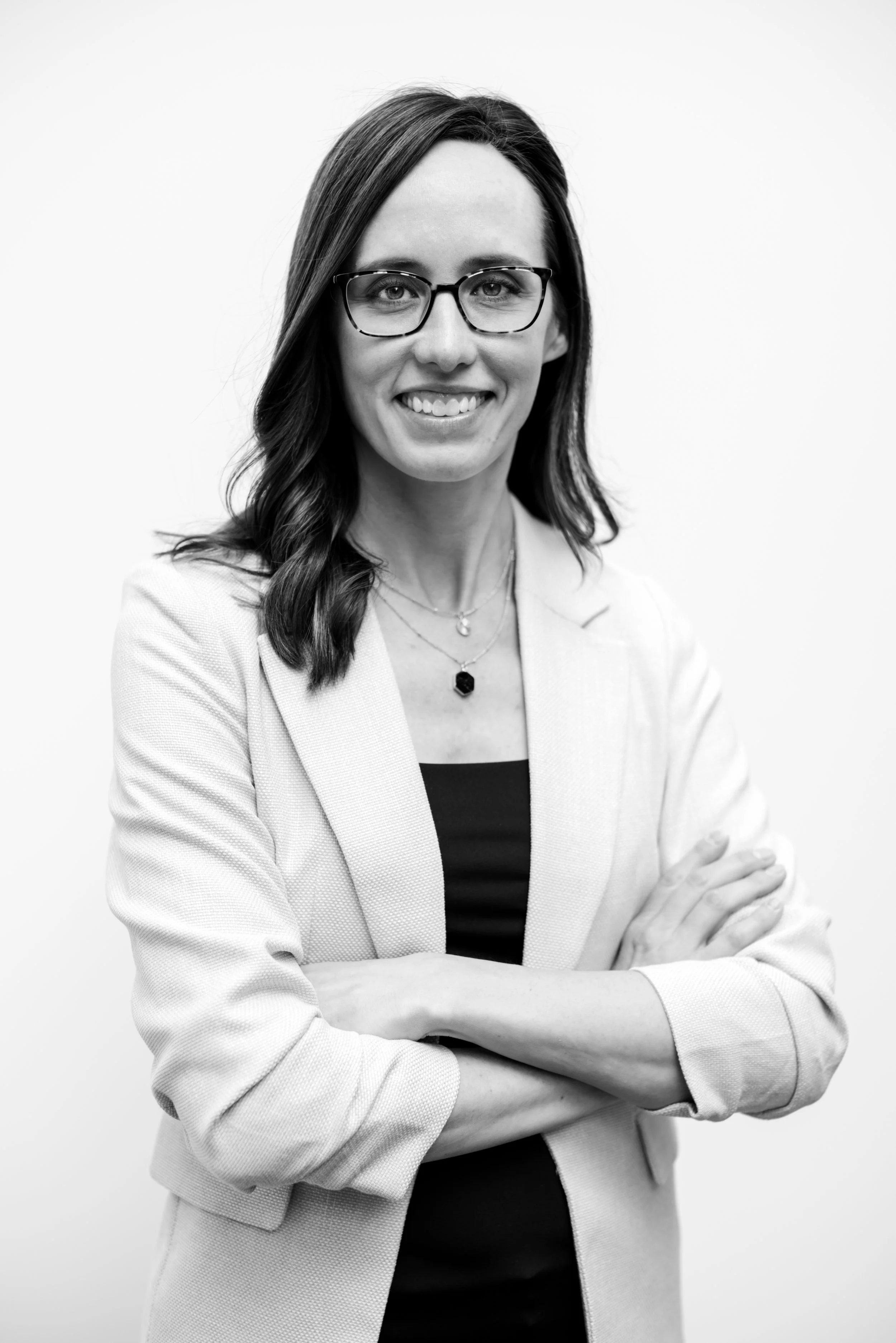 Black and white portrait of a woman with glasses and shoulder-length dark hair, smiling, wearing a light blazer over a dark top, with crossed arms.