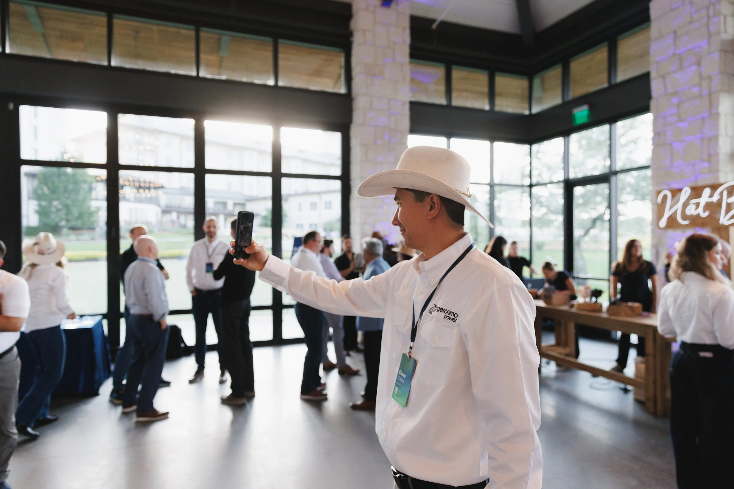 A man wearing a cowboy hat and white shirt taking a selfie at a business event in a modern, well-lit venue with large windows, other attendees, and a sign that reads "Nat" in the background.