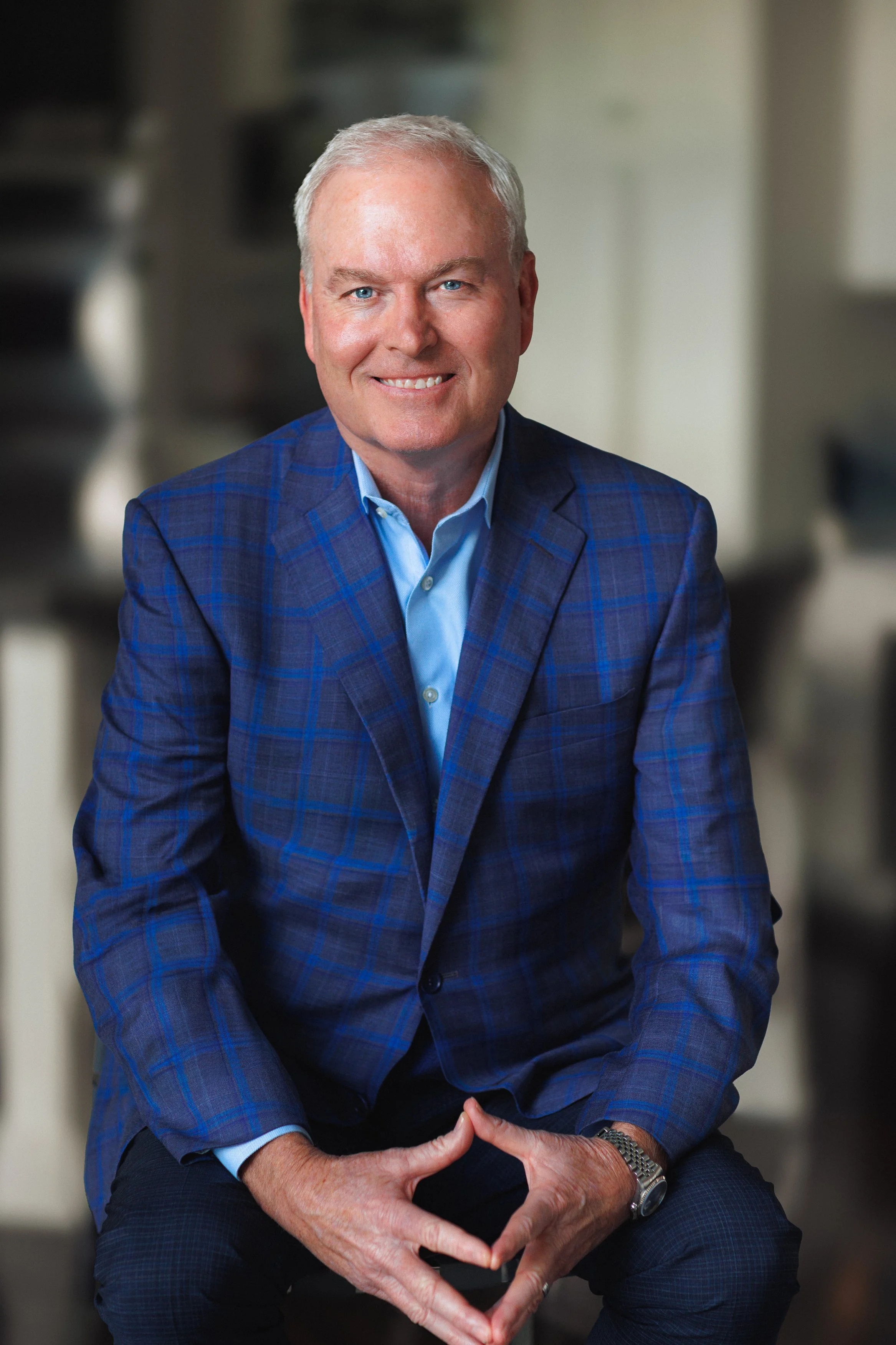 A man in a blue blazer and light blue shirt smiling, sitting with hands clasped, in an indoor setting with a blurred background.
