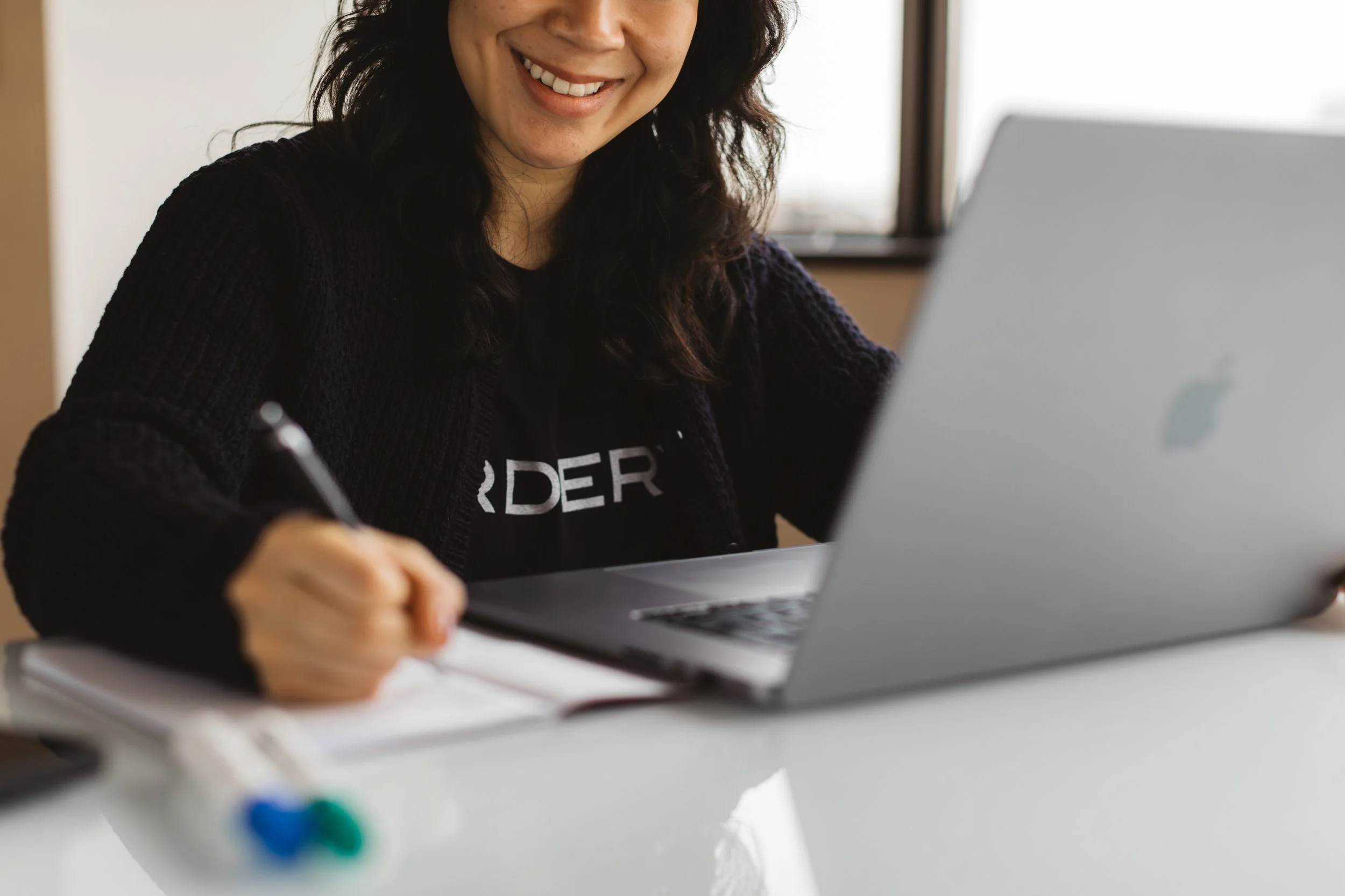 A woman smiling at a desk with a laptop, writing in a notebook, in a well-lit room.