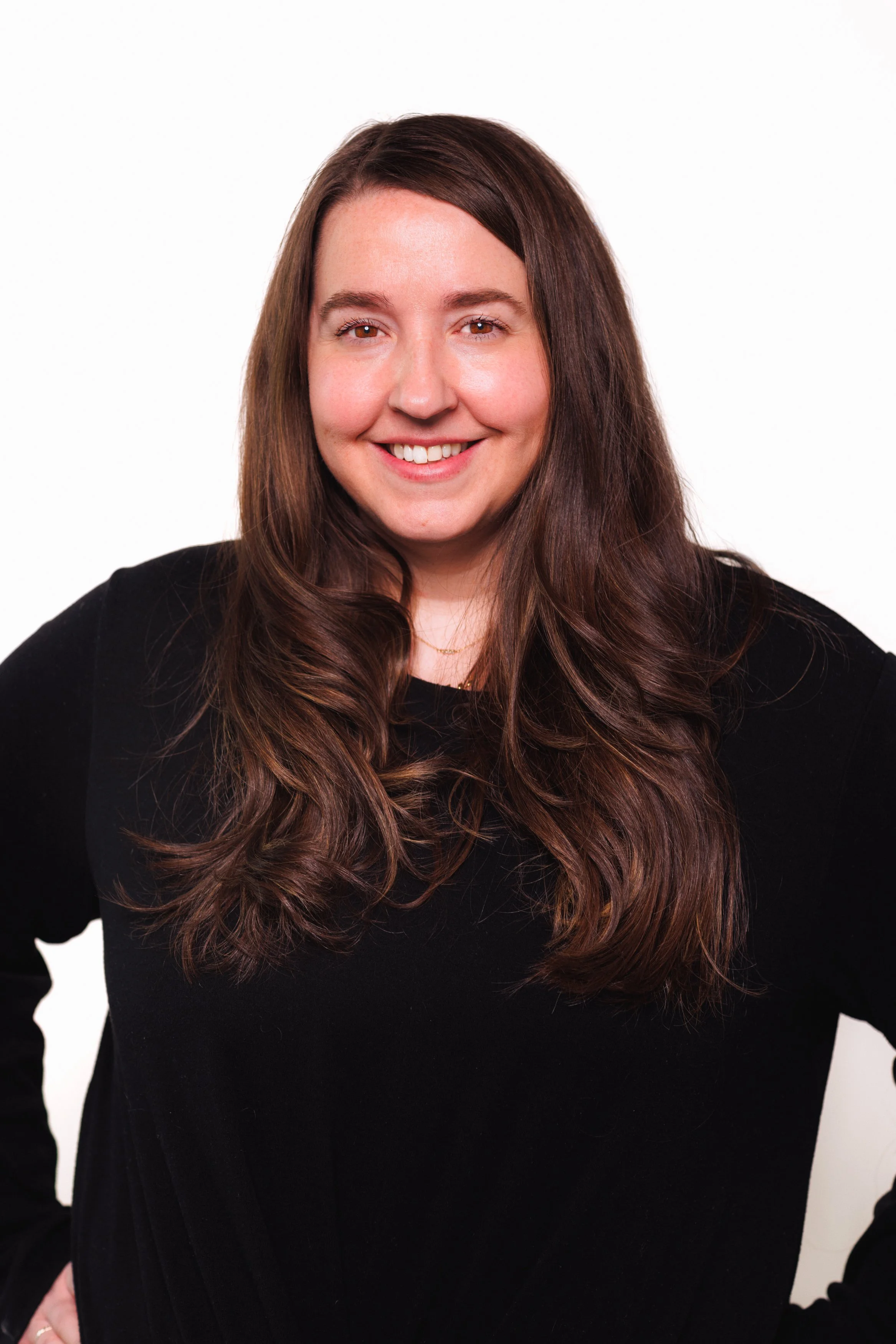 A woman with long brown hair, wearing a black top, smiling with hands on hips, against a white background.