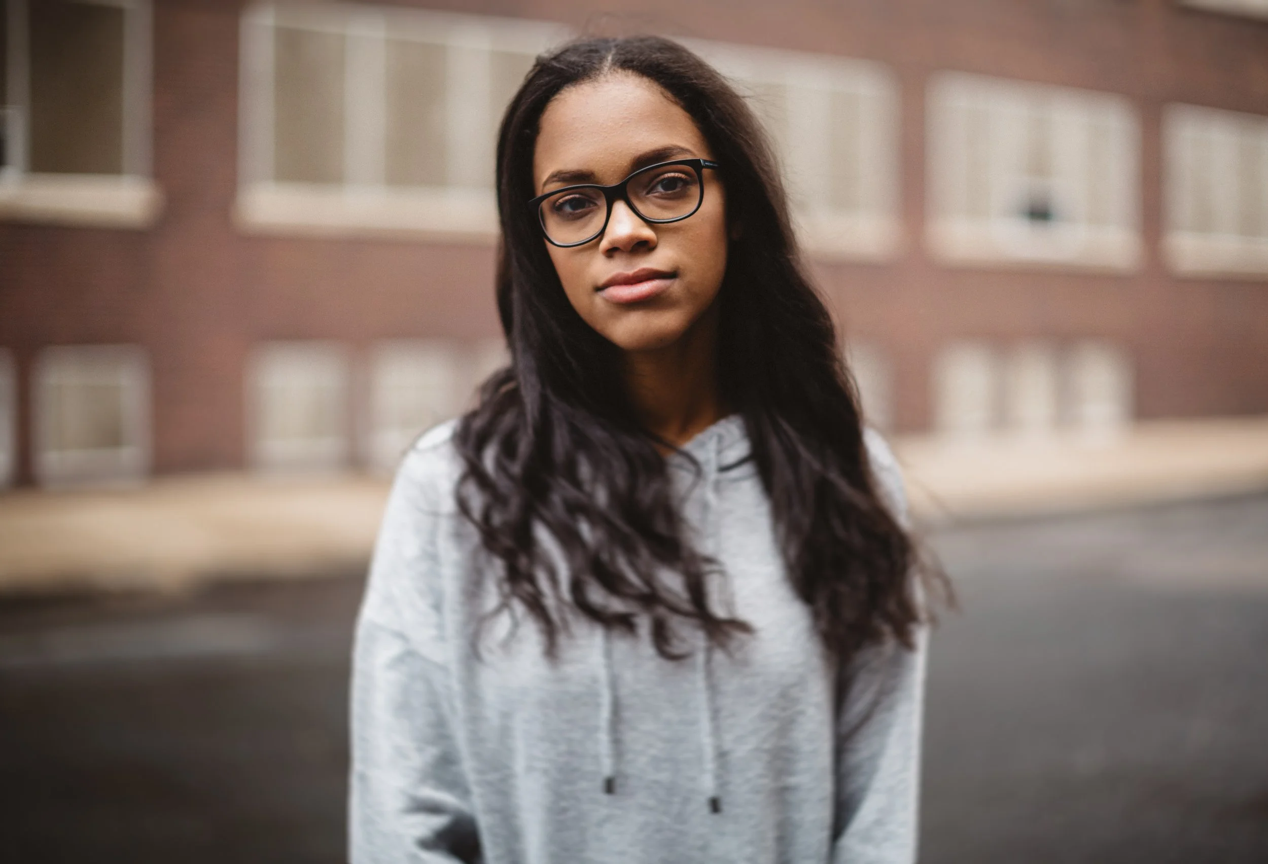 Young woman with glasses and long dark hair standing outdoors in front of a brick building, wearing a gray hoodie.
