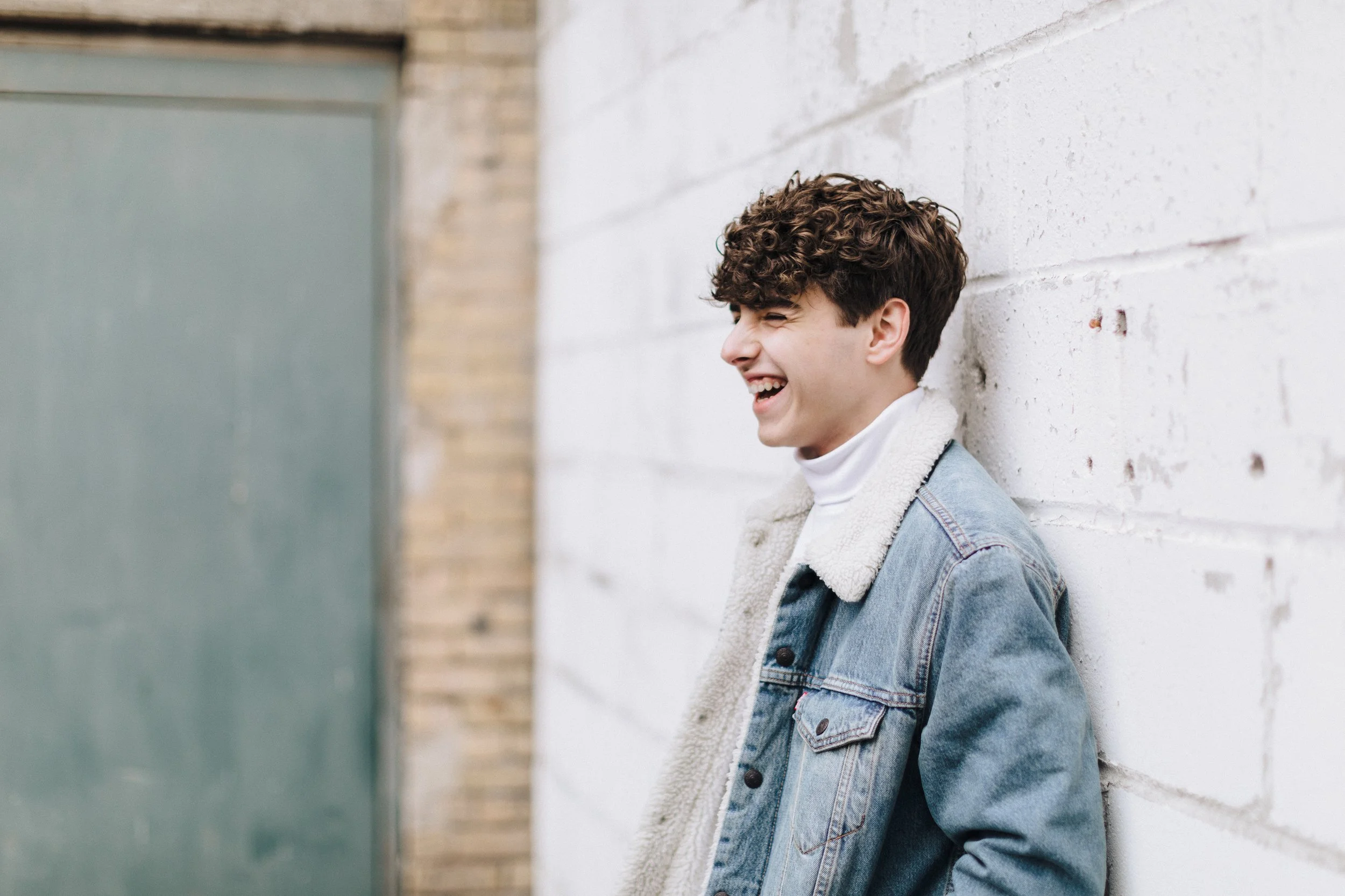 Young man with curly hair laughing while leaning against a white brick wall