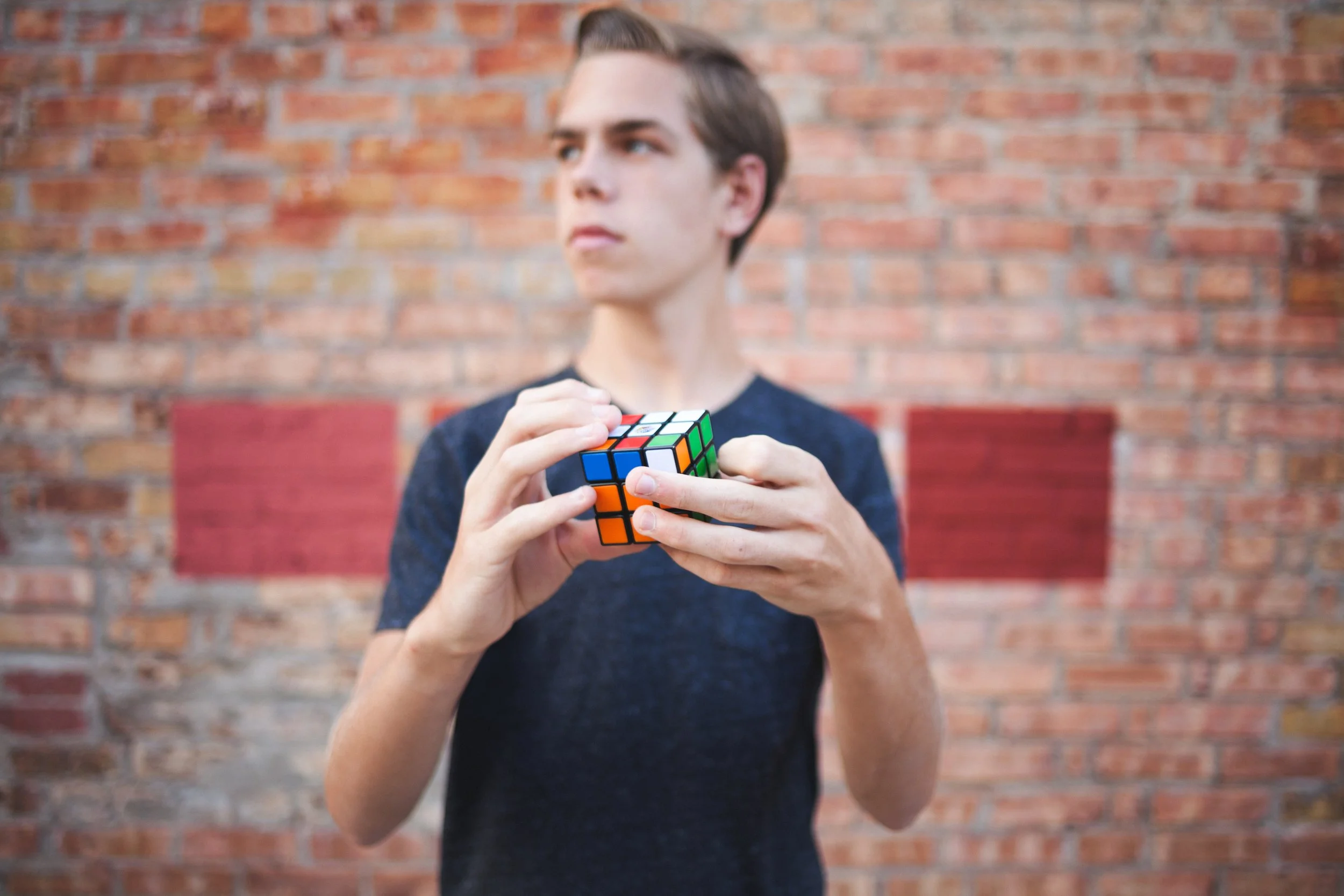 A young man with short hair stands in front of a brick wall, holding and looking at a solved Rubik's Cube.