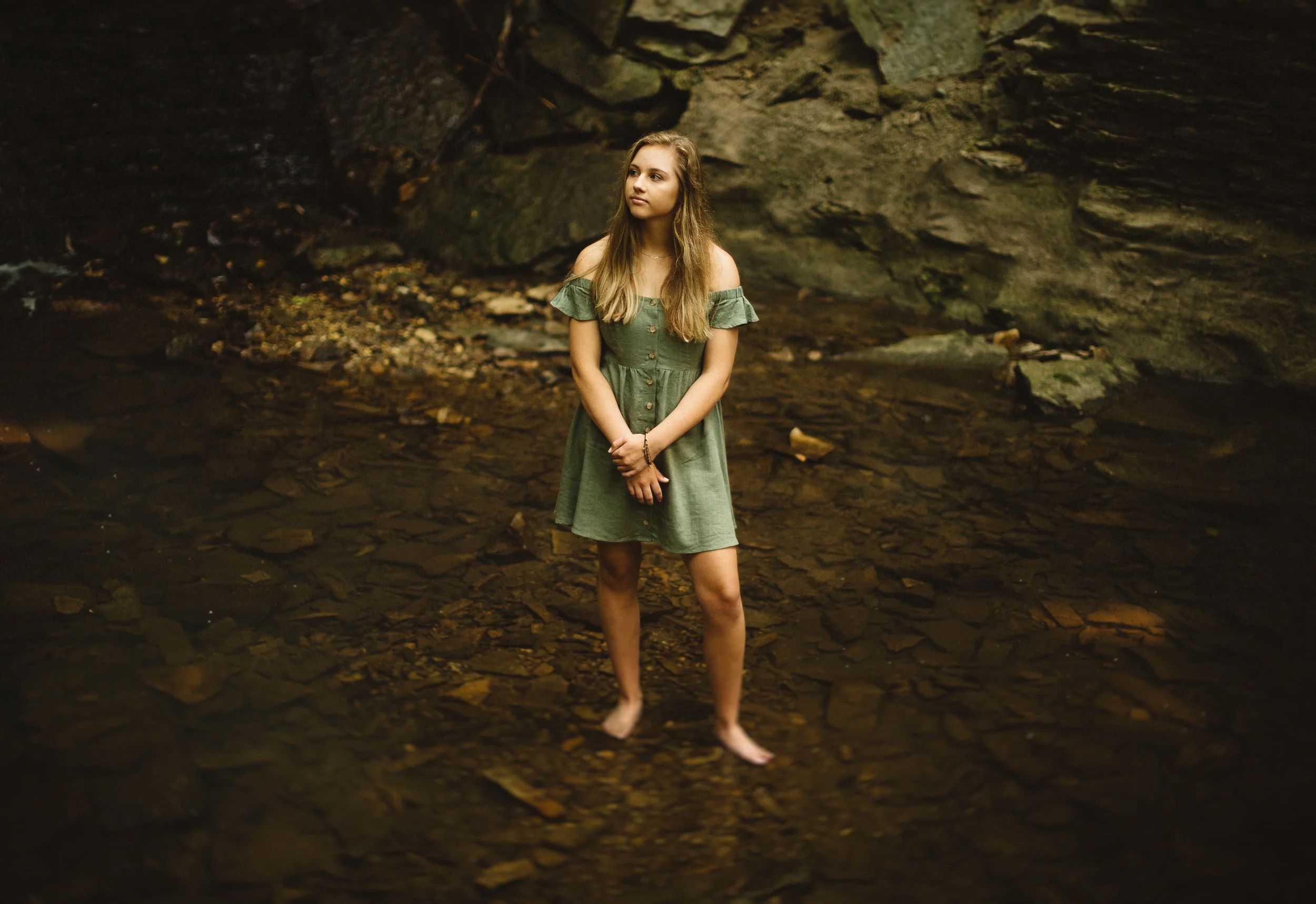 A young woman in a green dress standing barefoot in a shallow creek, with rocky terrain and a large rock face in the background.