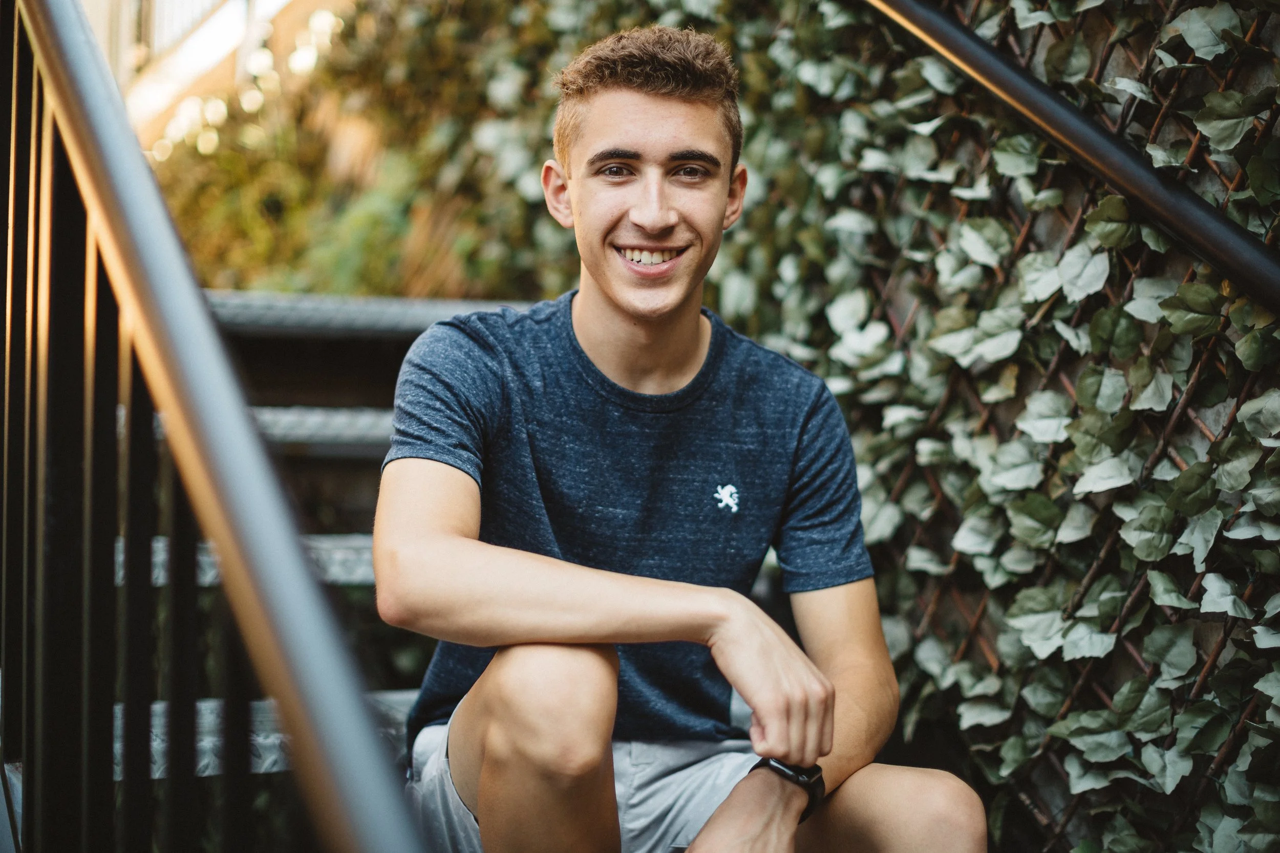 A young man with short curly hair smiling, sitting on outdoor stairs, wearing a dark blue t-shirt and light-colored shorts, with a background of green leaves and a black metal railing.