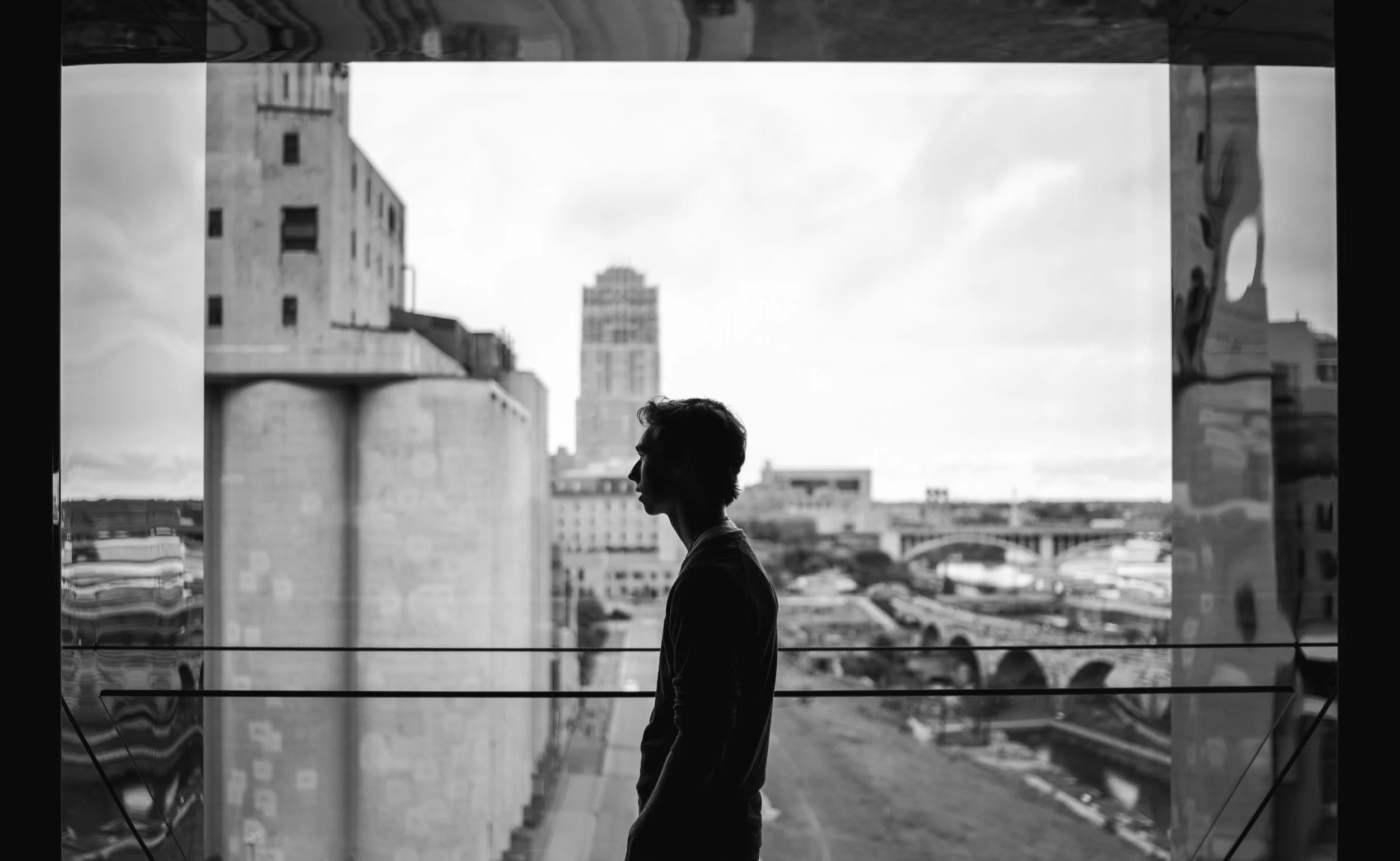 Silhouette of a person standing indoors near a large window with a cityscape background, black and white photo.