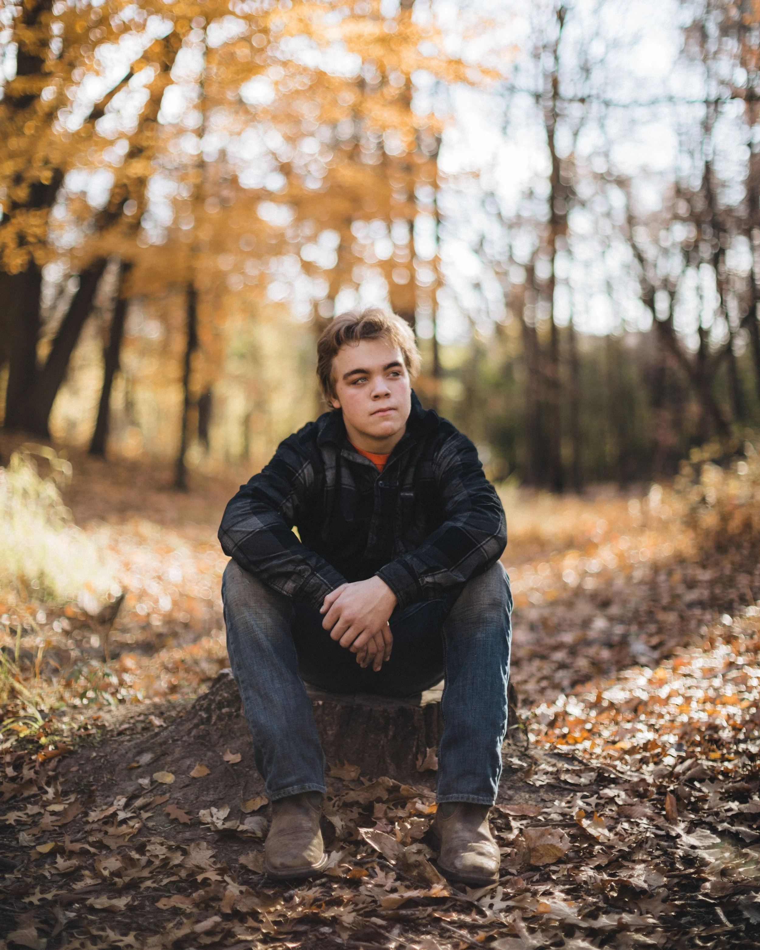 A teenage boy sitting on a tree stump in a fall forest with yellow and brown leaves, wearing a black plaid shirt, jeans, and boots.