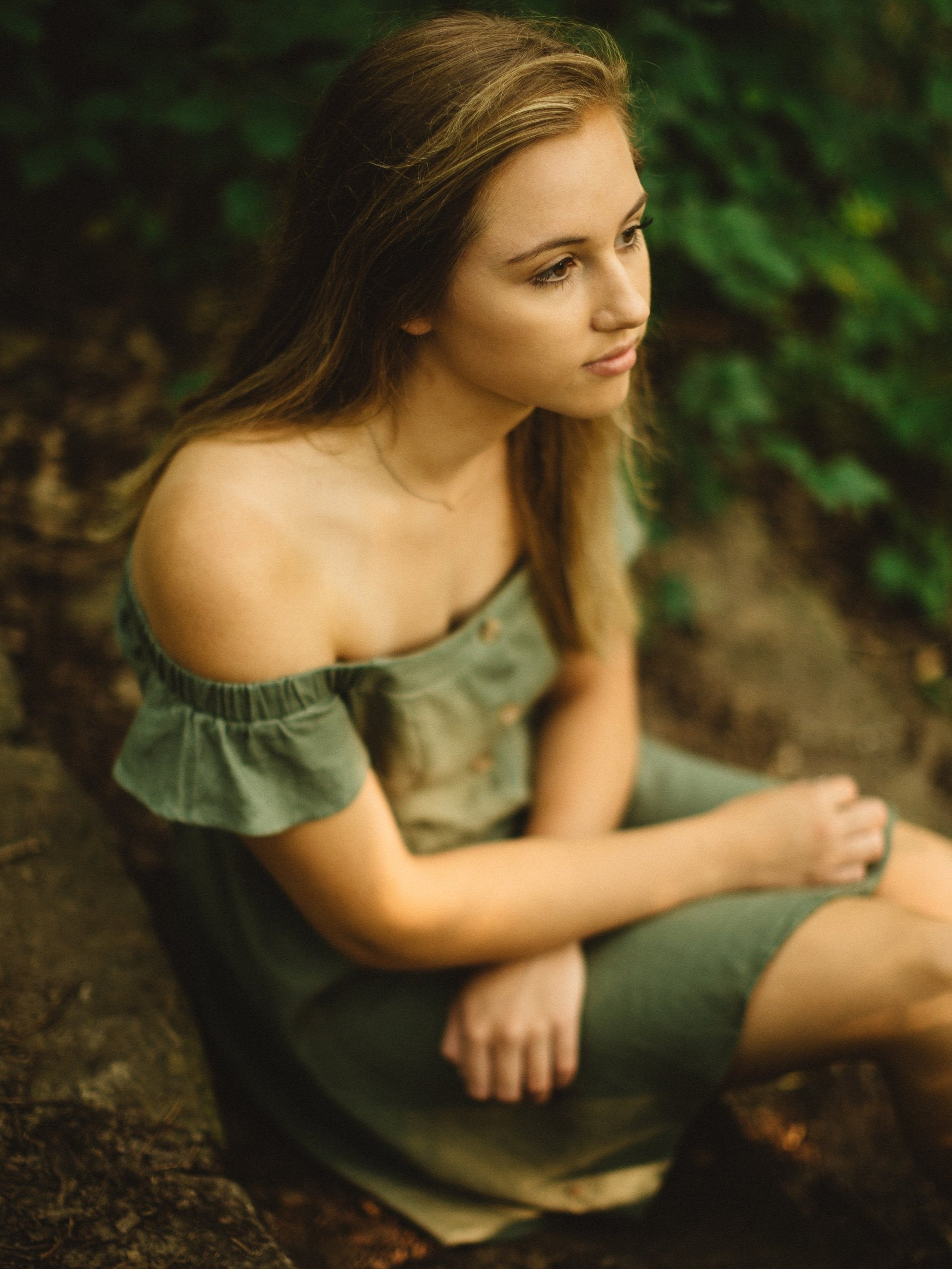 A young woman with long, light brown hair sitting outdoors in a natural setting, wearing an off-the-shoulder green dress, looking thoughtfully to the side.