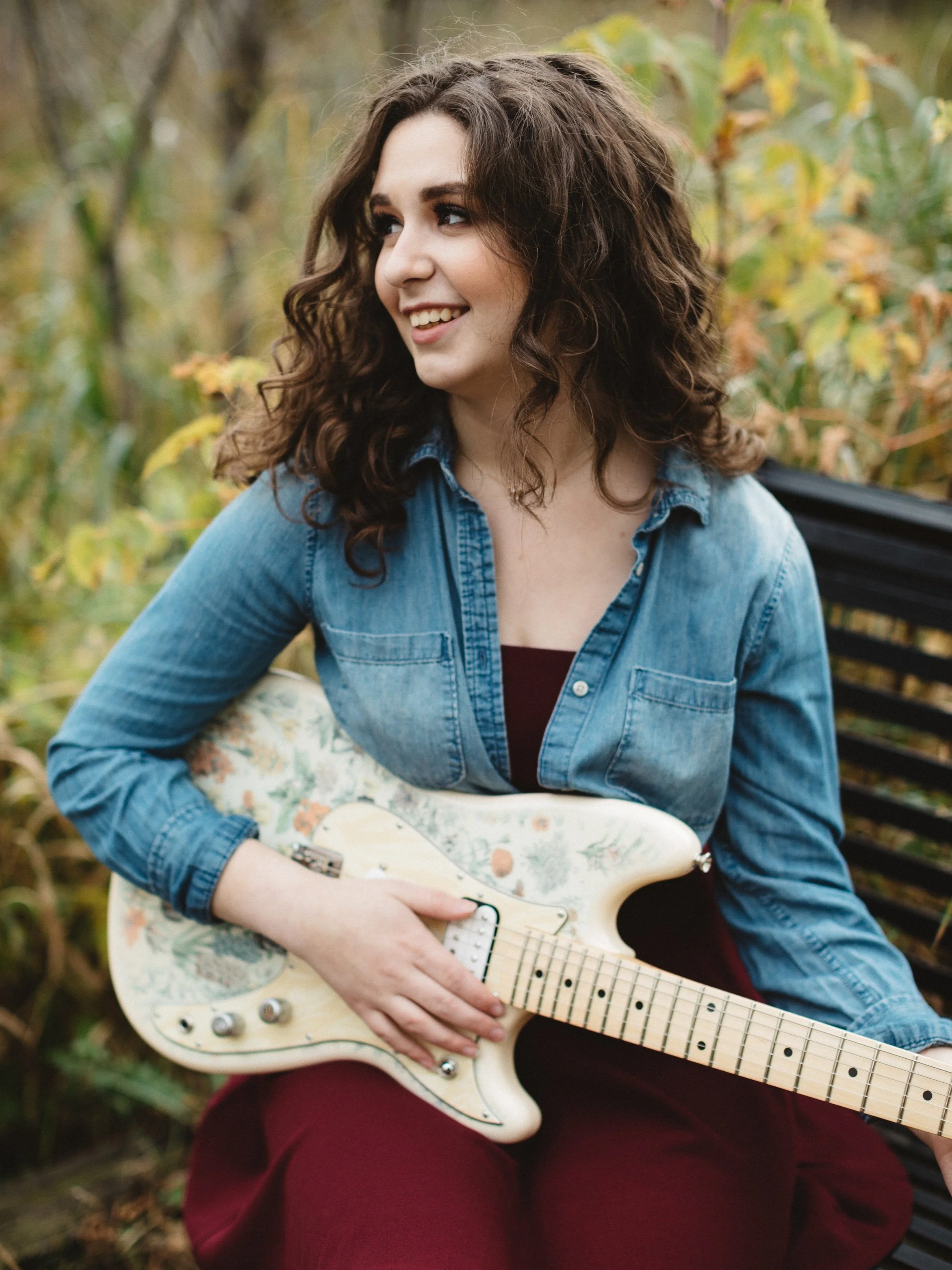A young woman with curly brown hair, wearing a denim shirt and maroon pants, sitting on a park bench outdoors with autumn foliage in the background, holding an electric guitar and smiling.