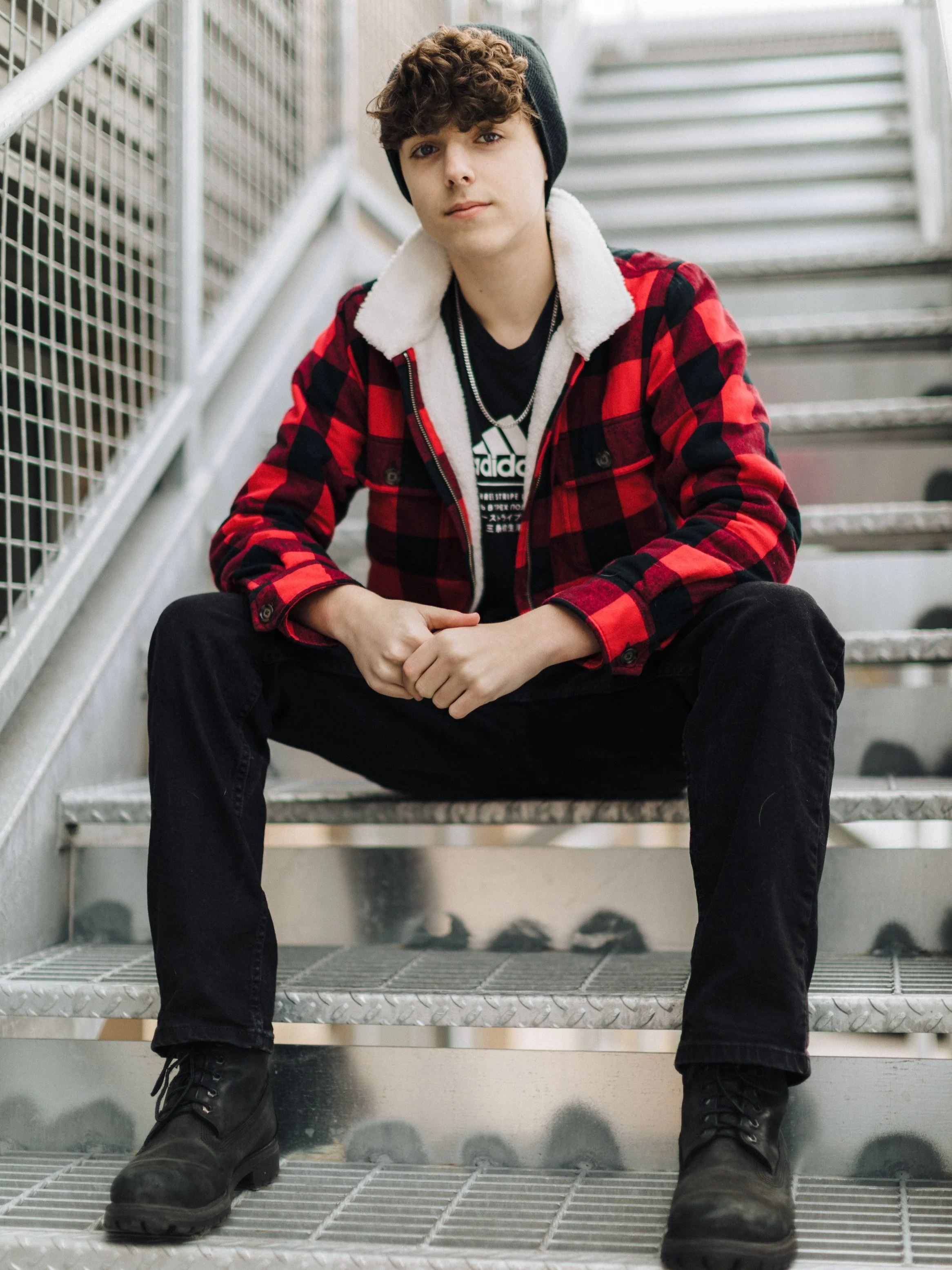 A young man sitting on outdoor stairs, wearing a black beanie, red and black plaid jacket with a white fleece collar, black jeans, and black boots, looking at the camera.