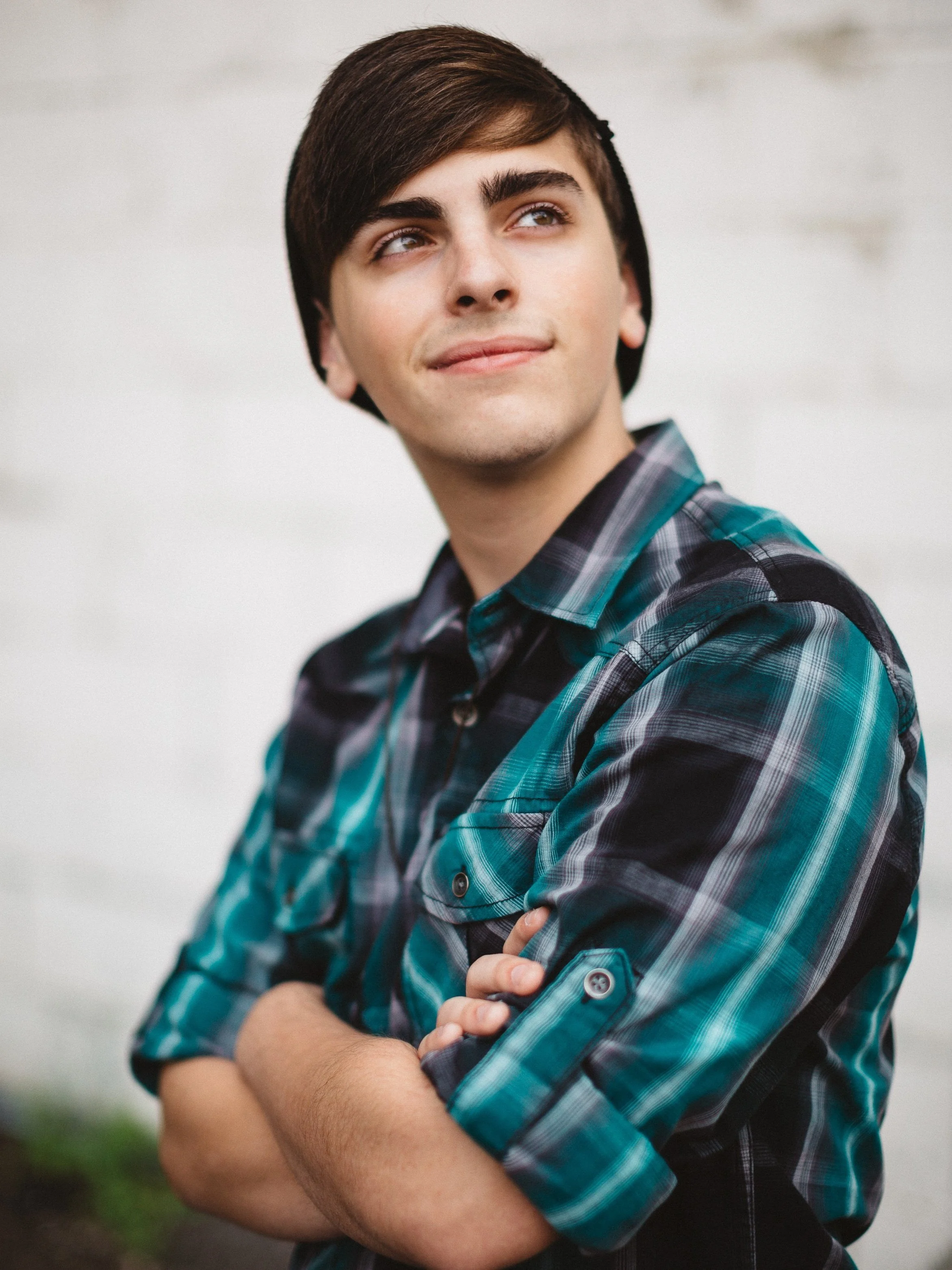 A young man with dark hair, wearing a black beanie and plaid shirt, smiling slightly and crossing his arms, standing outdoors against a light background.