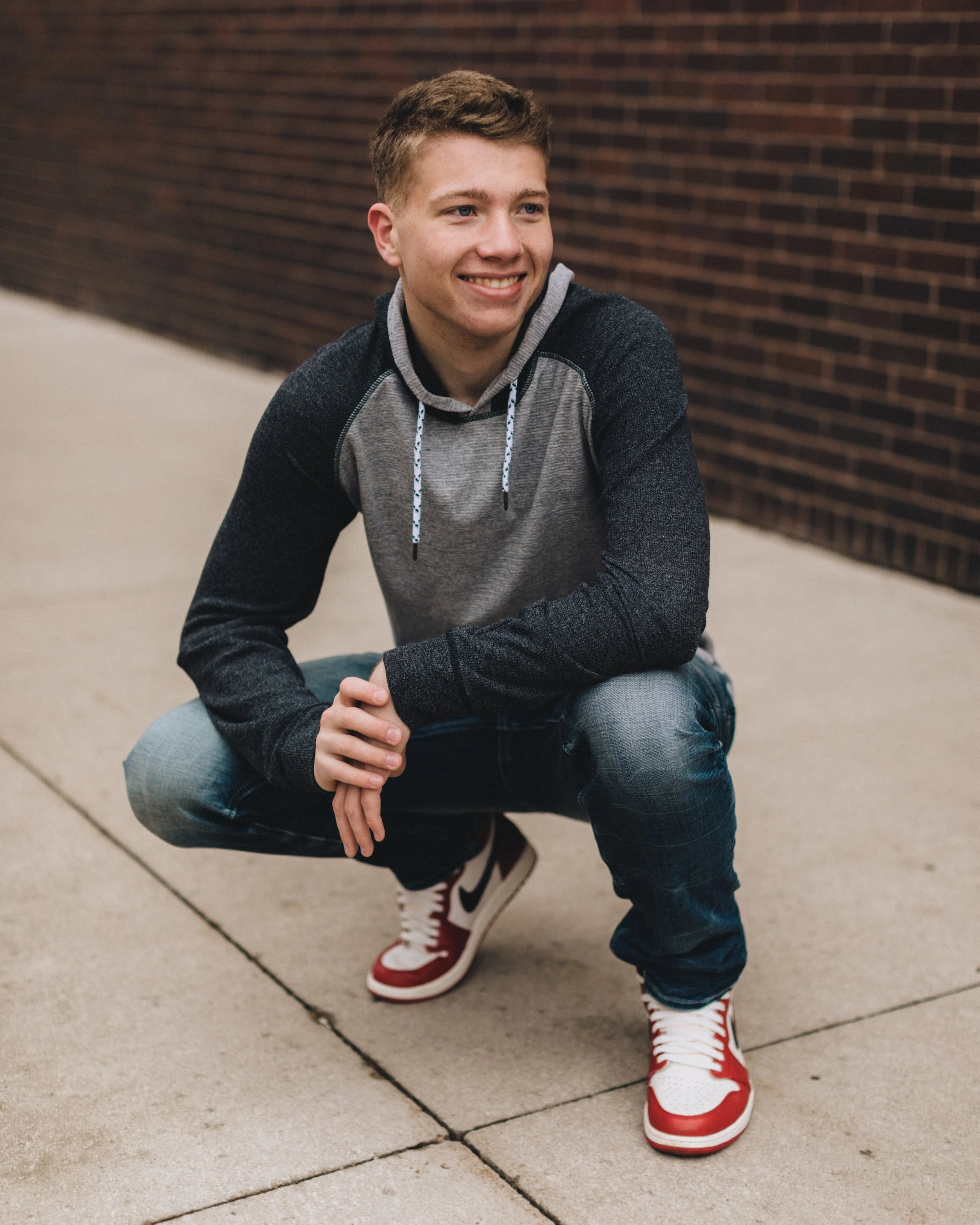 Young man squatting on sidewalk next to brick wall, smiling, wearing gray and black hoodie, blue jeans, and red and white sneakers.