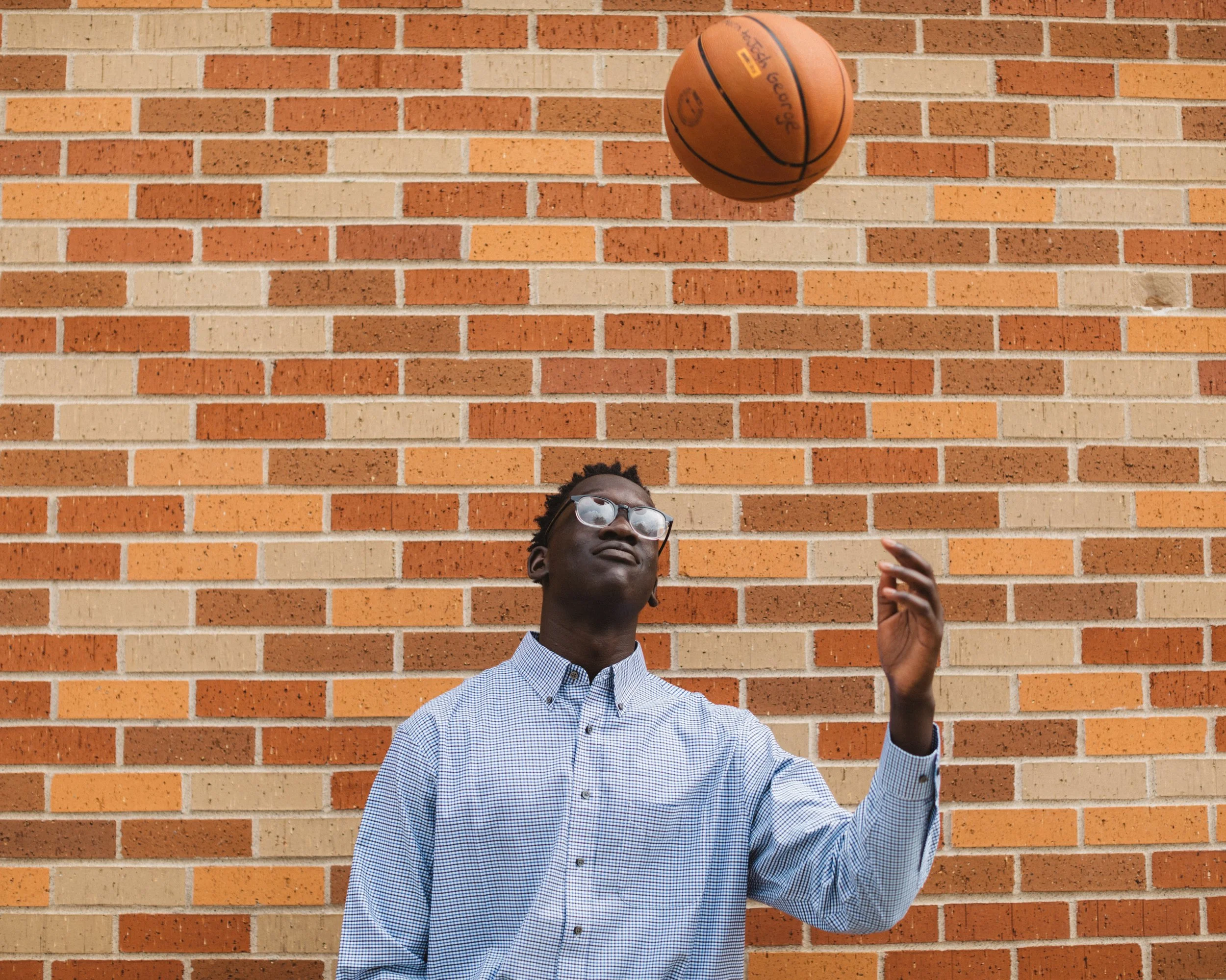A man wearing glasses and a checkered shirt standing in front of a brick wall, playing with a basketball.