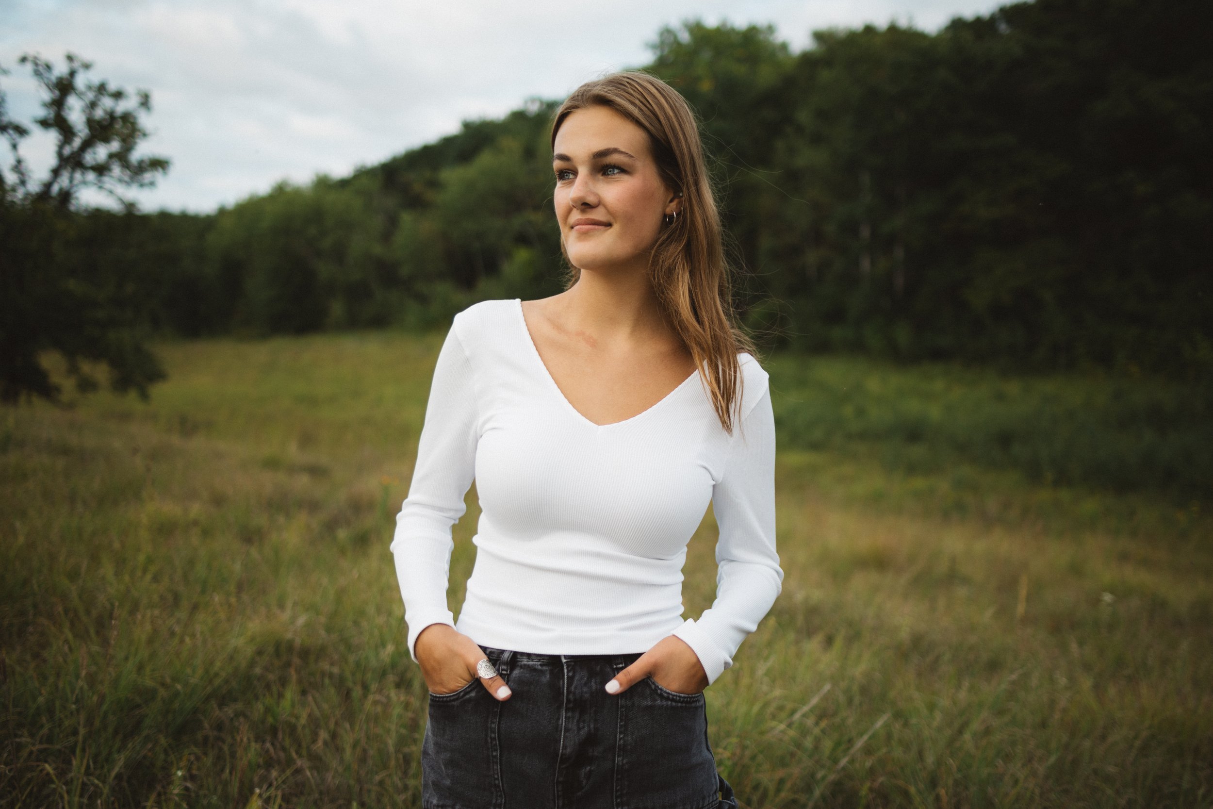 Young woman standing outdoors in a grassy field with trees in the background, wearing a white long-sleeve top and black shorts.
