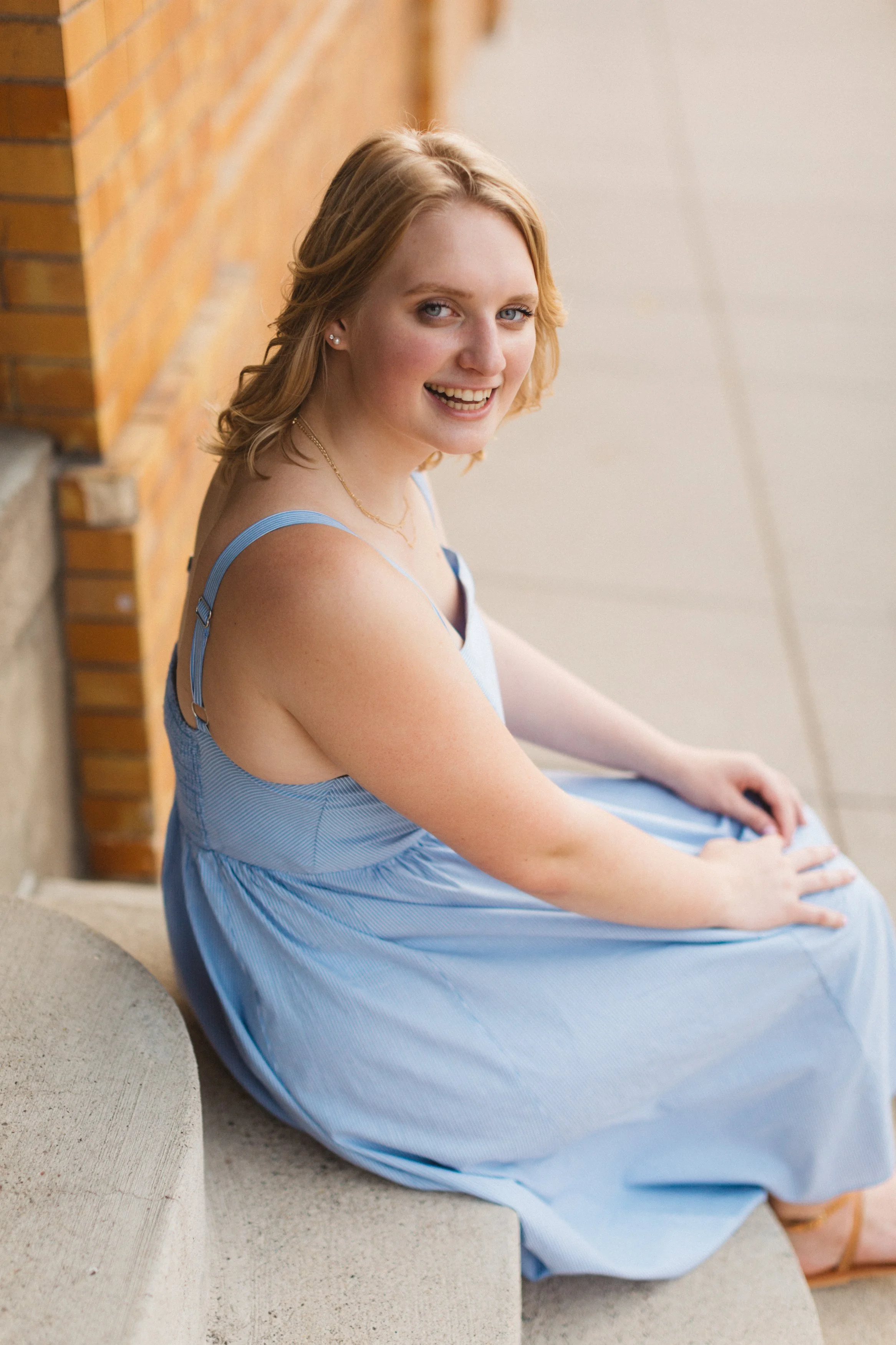 A young woman with shoulder-length blonde hair sitting on a curved concrete surface, wearing a light blue dress, smiling at the camera, with a brick wall behind her.