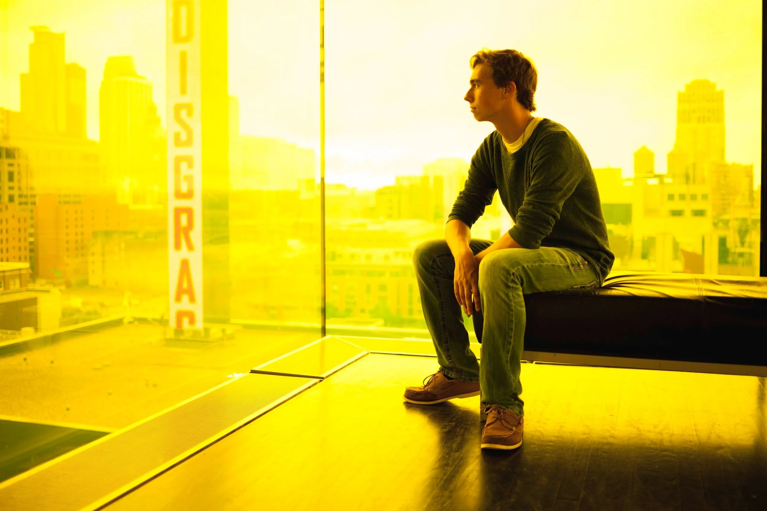 A young man sitting on the edge of a window seat, looking out at a cityscape with a yellow filter overlay.