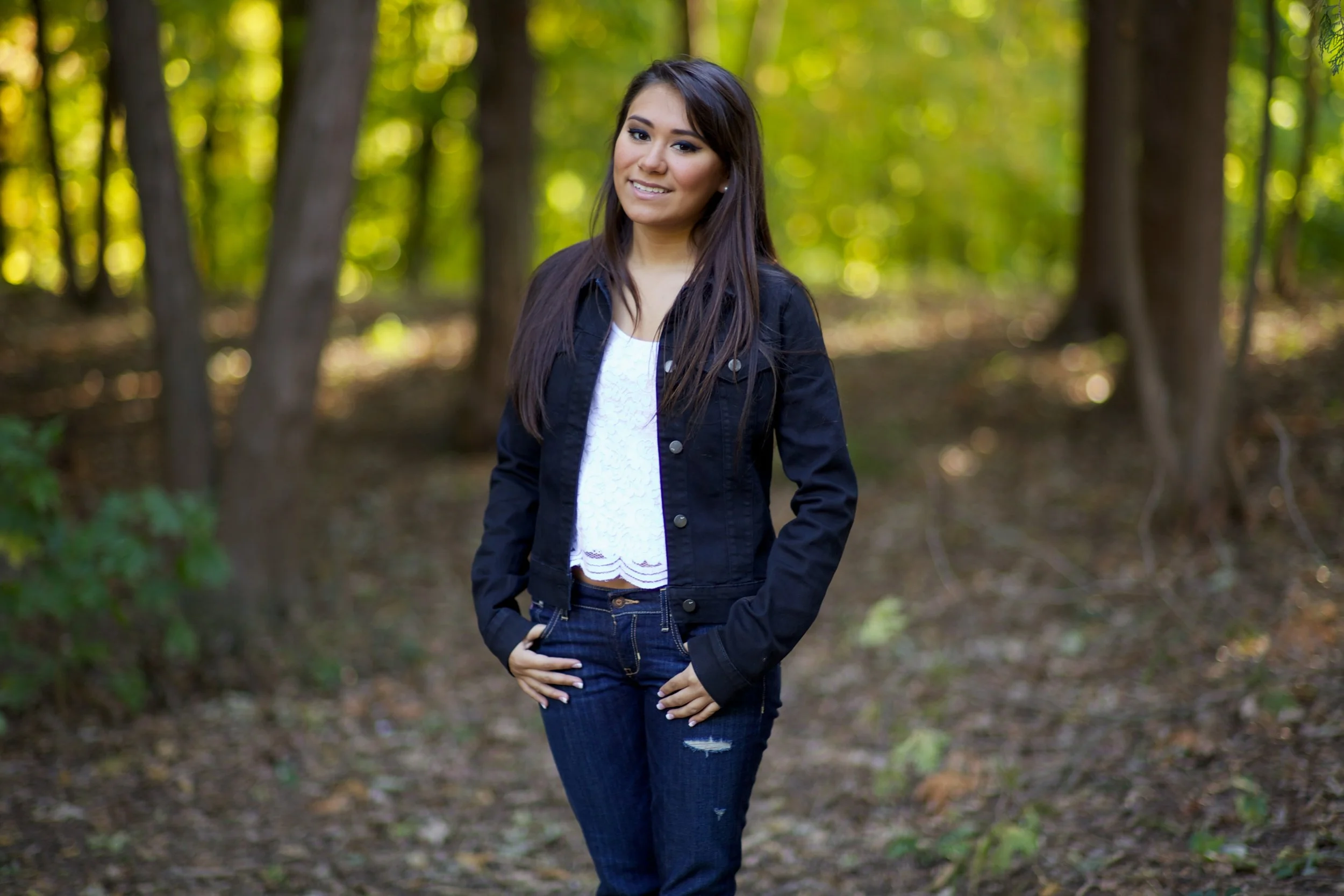 A young woman with long dark hair smiling and standing in a forest with trees and greenery in the background. She is wearing a black denim jacket, a white top, and ripped jeans.
