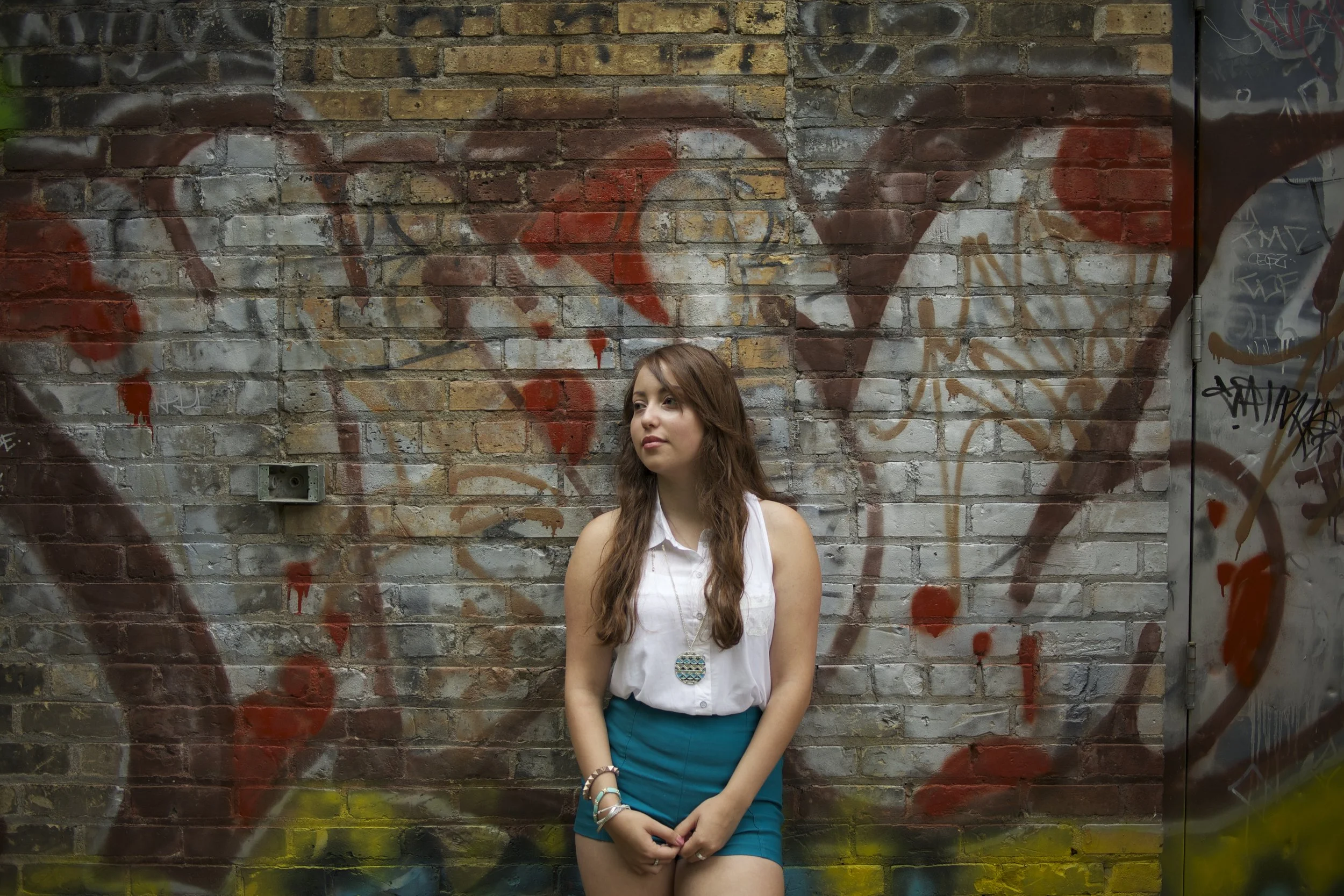 A young woman with long wavy brown hair stands against a graffiti-covered brick wall, wearing a white sleeveless blouse, teal shorts, and beaded jewelry, looking slightly to her left.