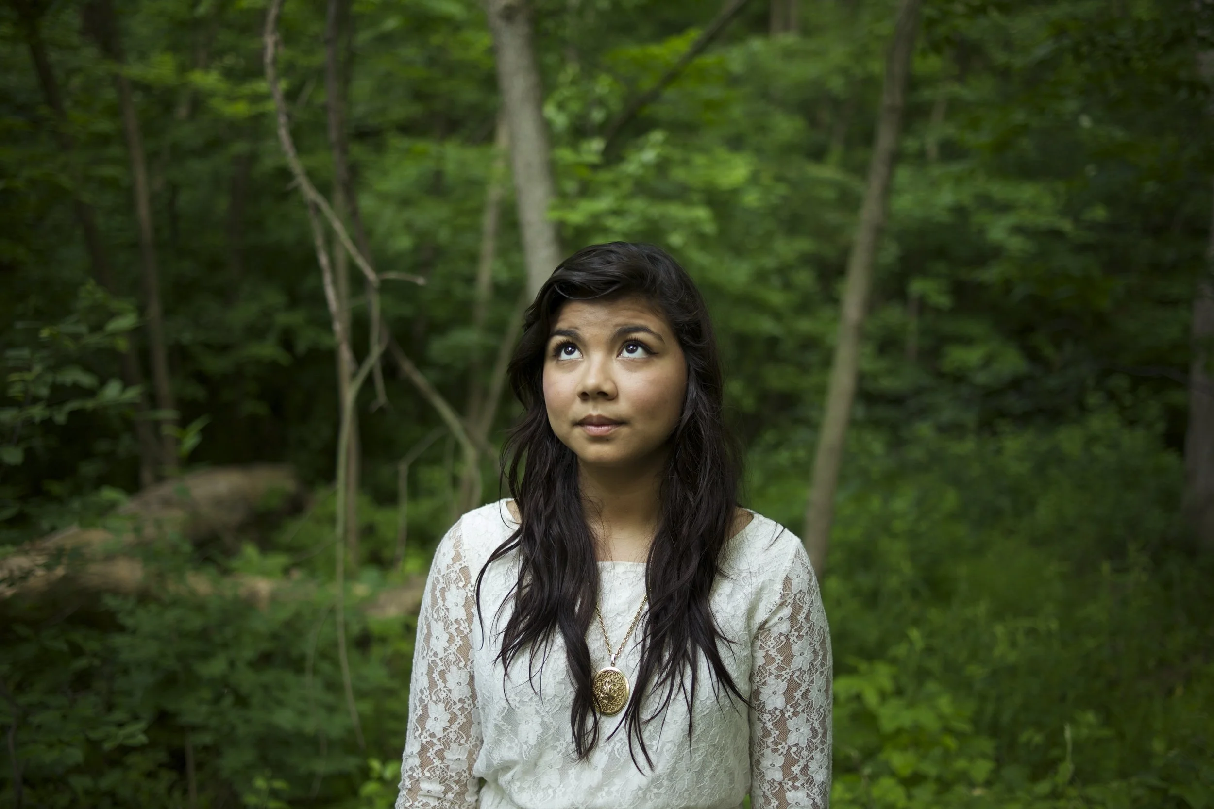 A young woman with dark hair wearing a white lace top and a gold medallion necklace standing in a green forest.