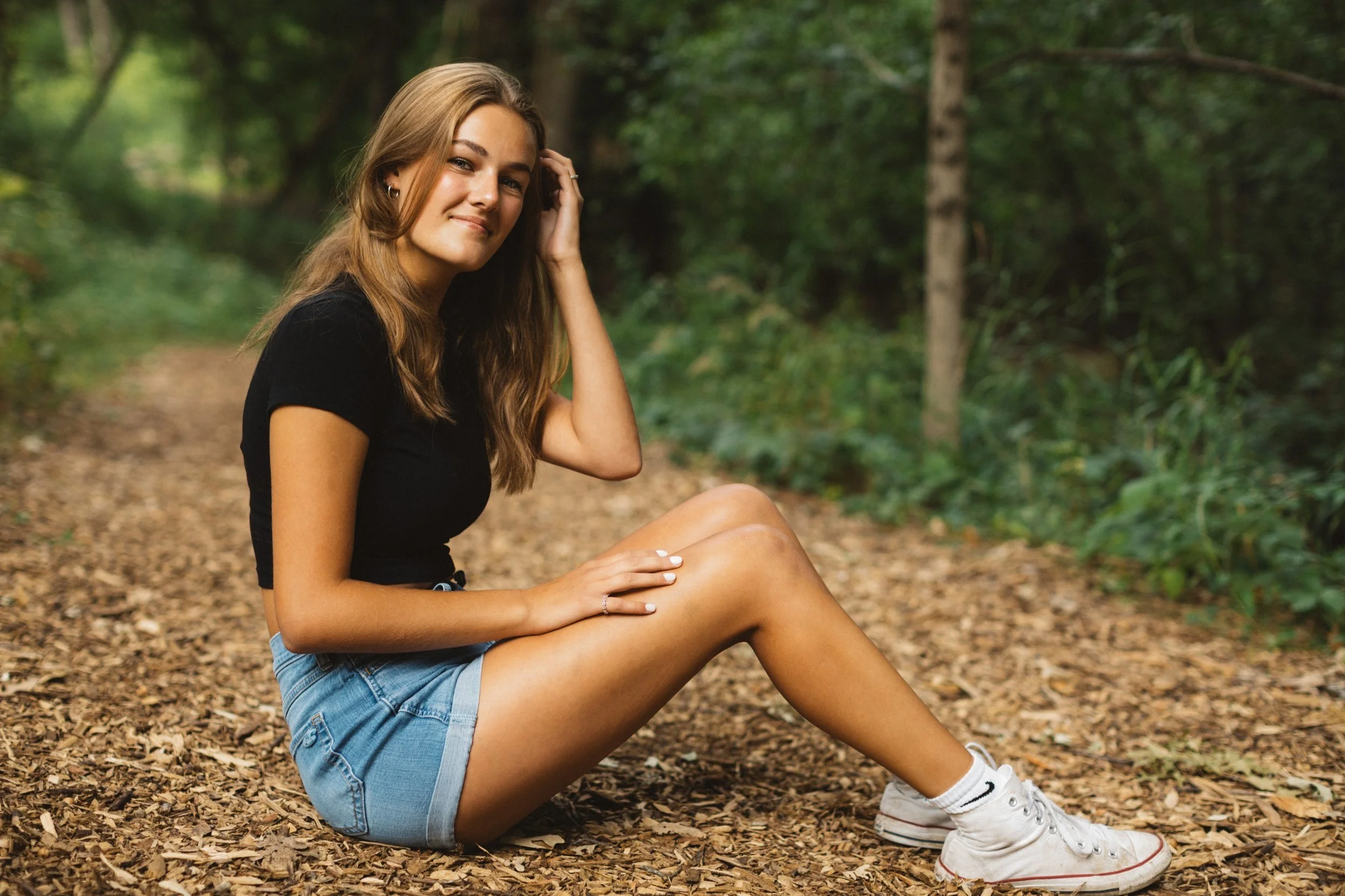 A young woman sitting on a leaf-covered path in a forest, wearing a black t-shirt, denim shorts, and white sneakers, smiling at the camera with her hand touching her hair.