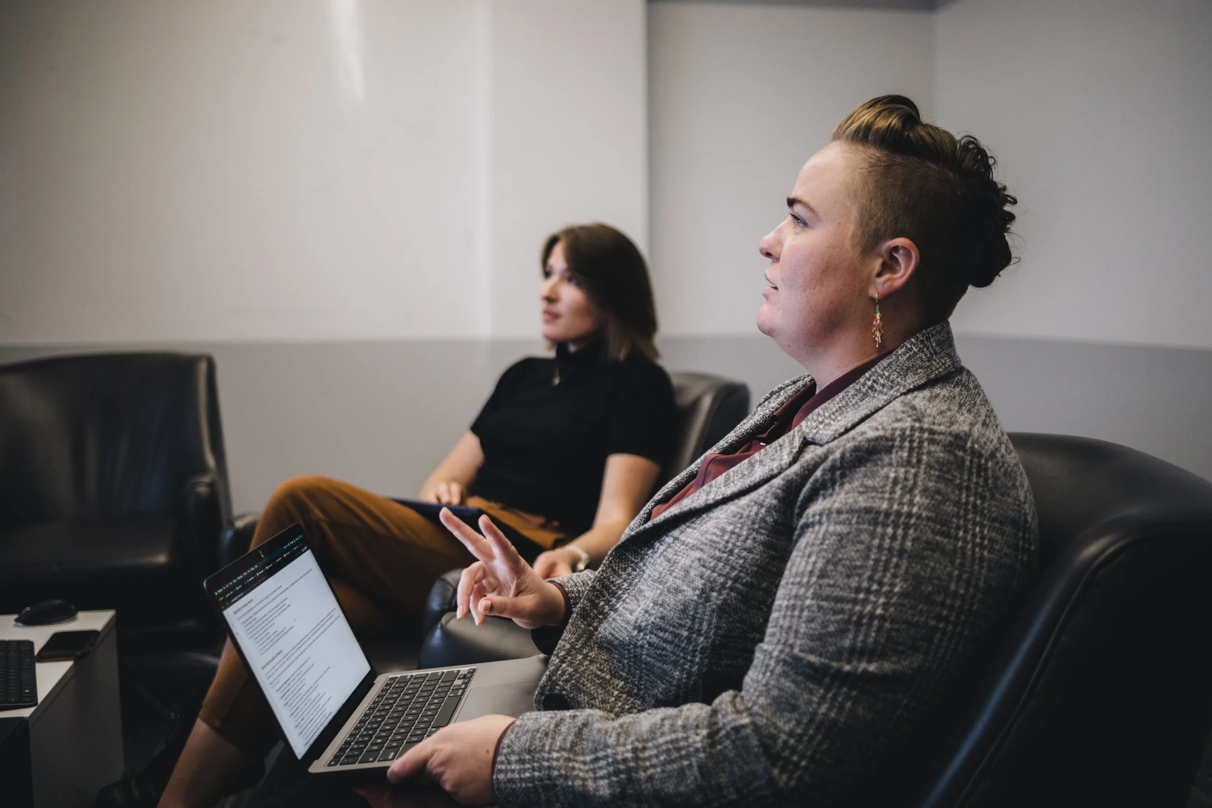 Two women seated in a meeting room, one is talking with a laptop on her lap, the other is blurred in the background, listening.