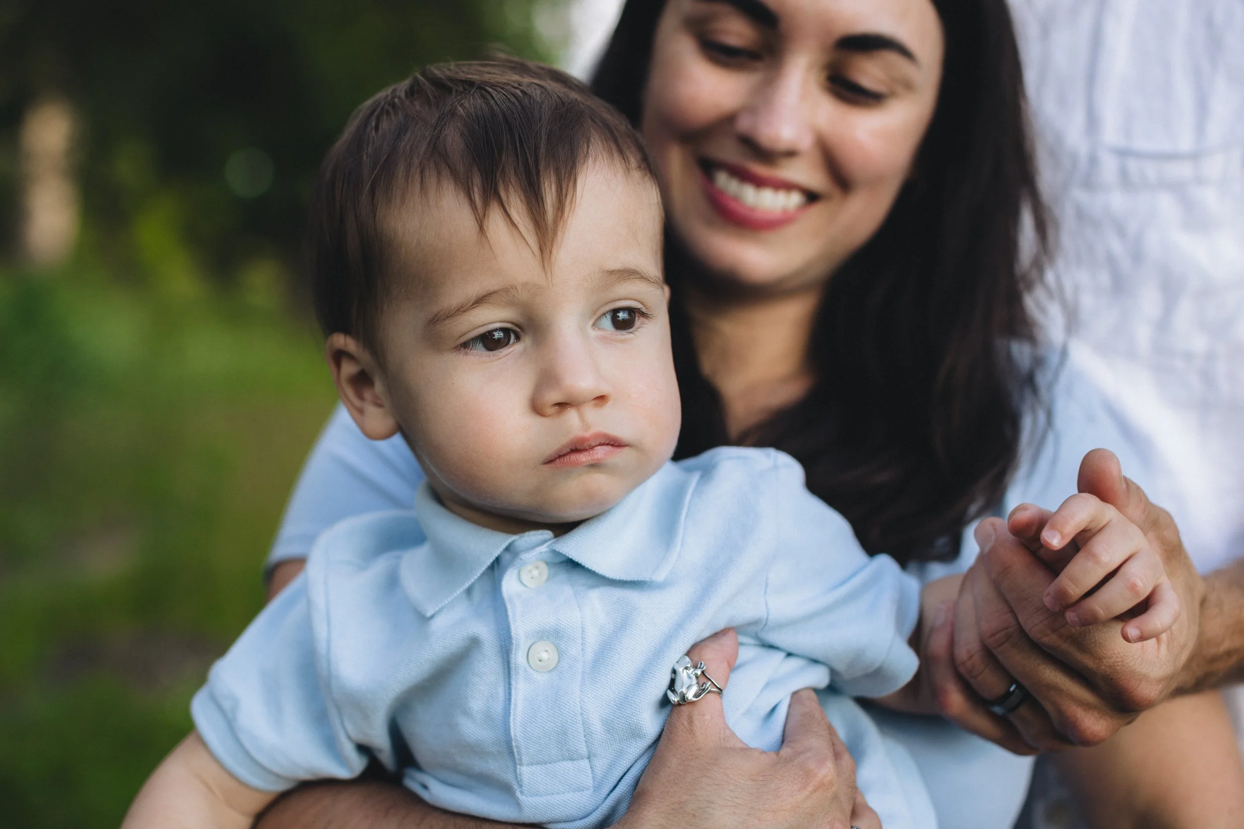 A woman holding a young boy outdoors. The boy has a serious expression, wearing a light blue shirt, while the woman smiles gently behind him.