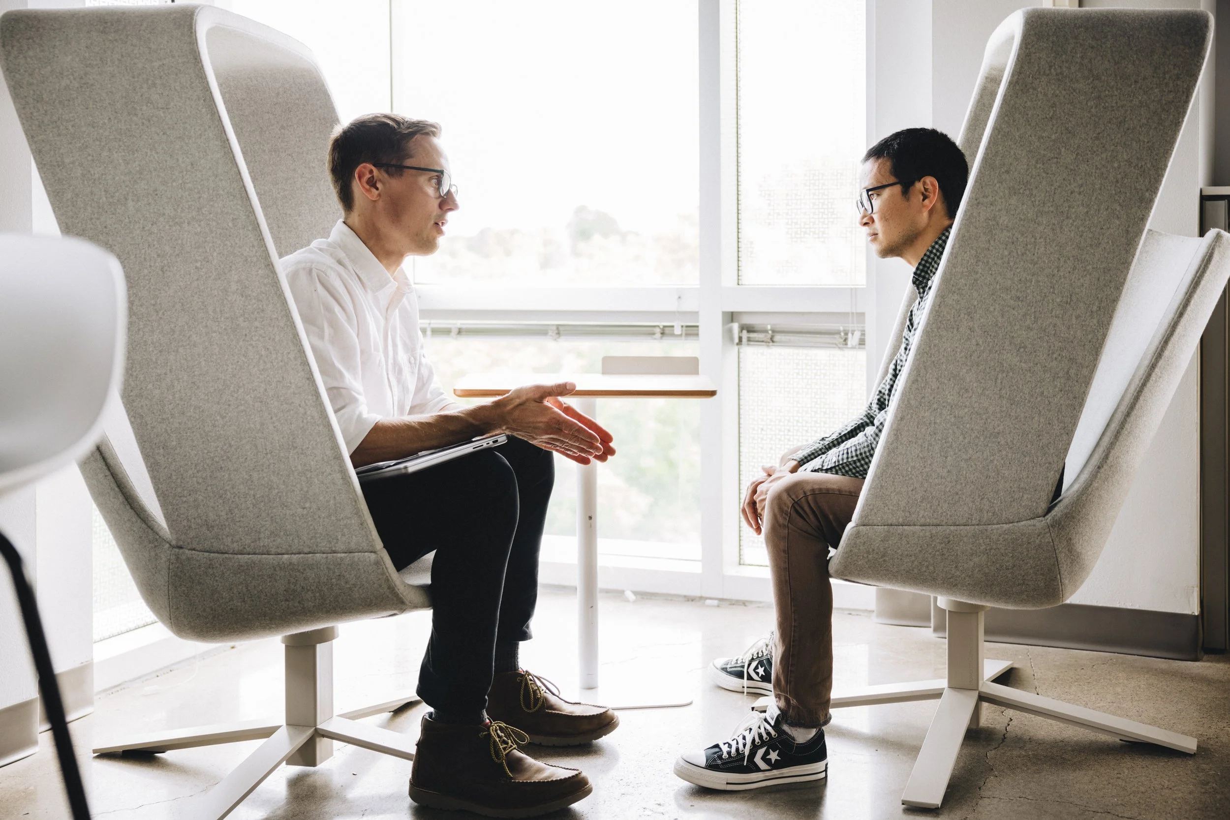 Two men sit facing each other in modern office chairs, engaged in a serious conversation, with a table between them, in a bright office with large windows.