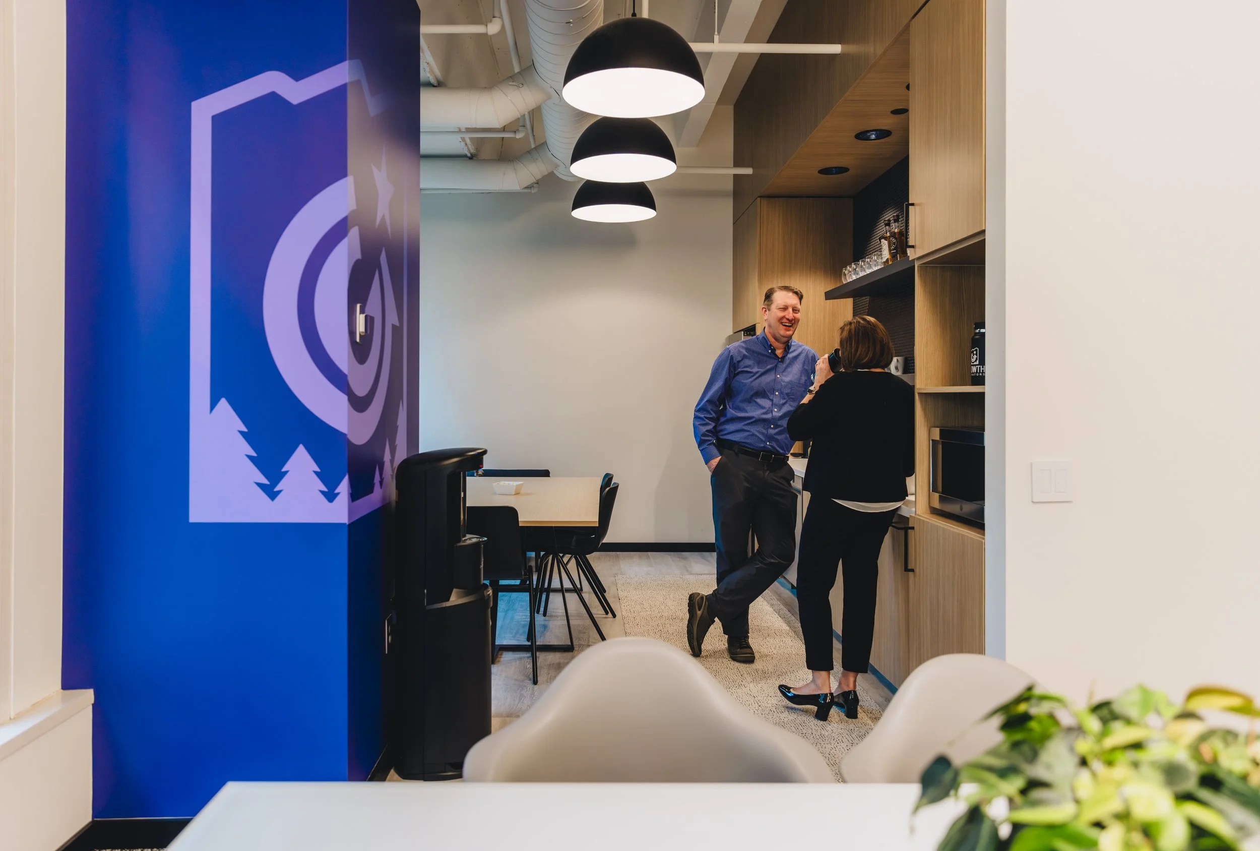 Two people are standing and talking in an office kitchen area, with a man laughing and wearing a blue shirt, and a woman facing away, holding a cup. The background includes a wooden cabinet, a microwave, and the ceiling has exposed ductwork and three