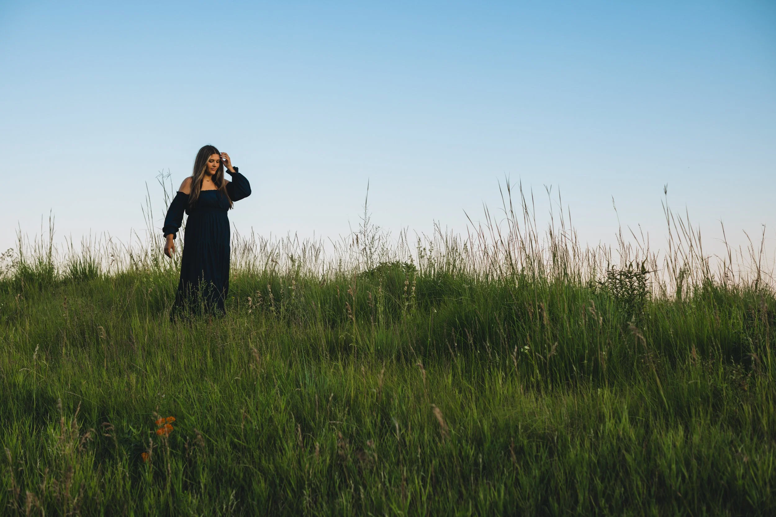 A woman in a black dress walking through tall green grass in a field during daytime with a clear blue sky in the background.