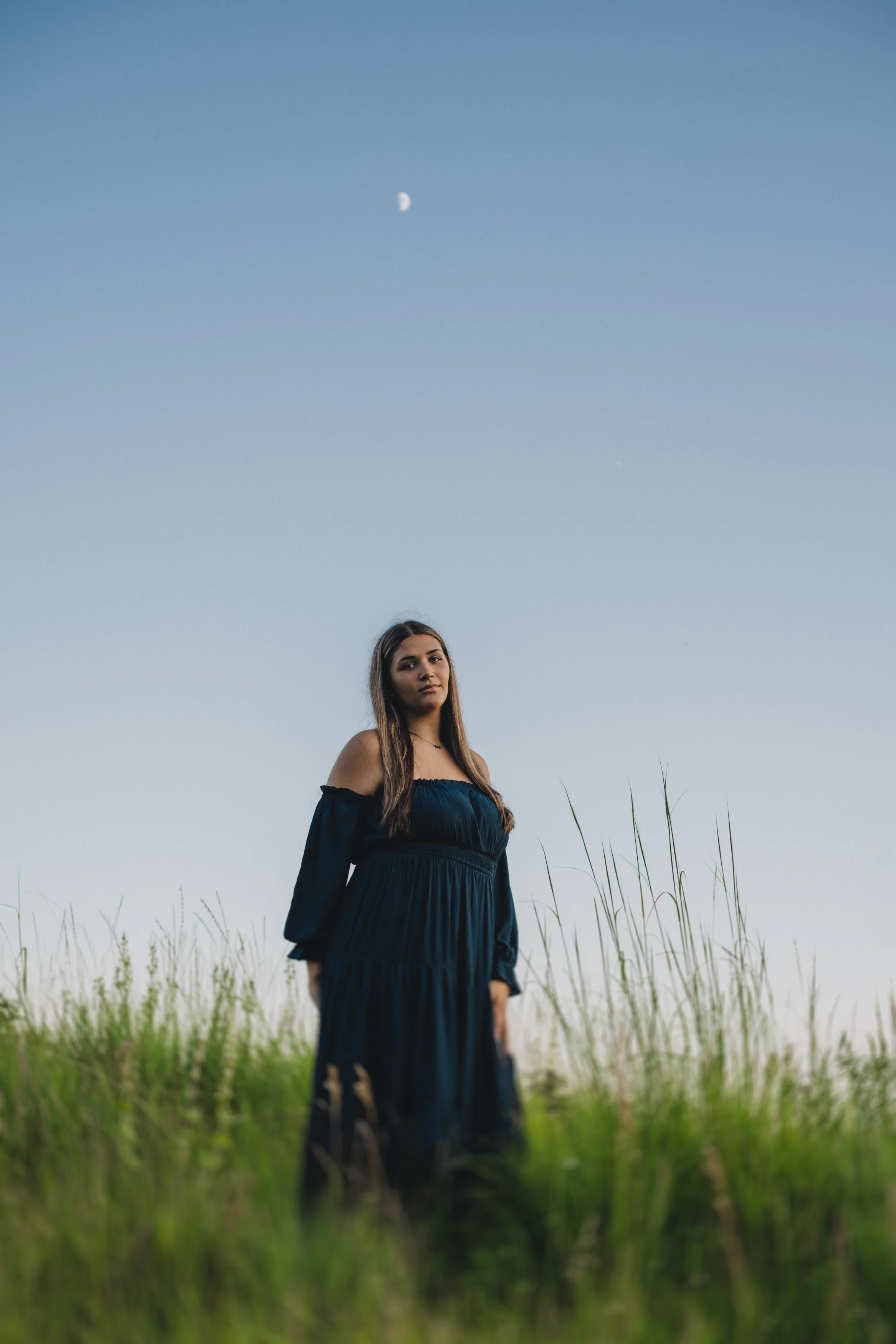 A young woman in a dark blue dress standing in a grassy field at dusk, with the moon visible in the sky.