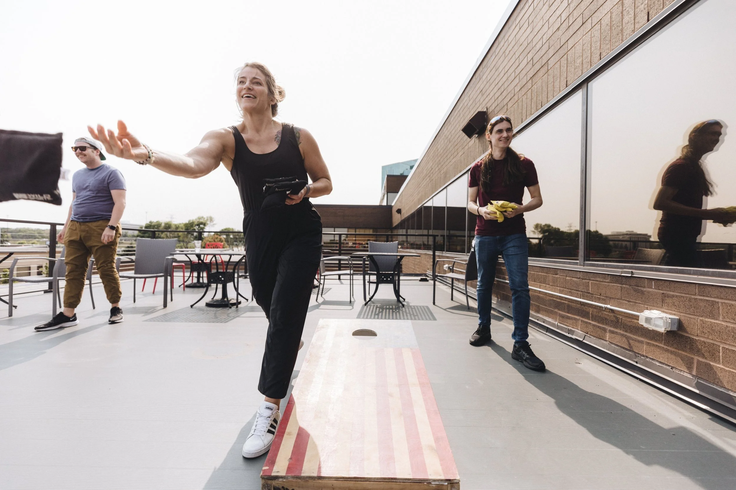 Three friends playing cornhole on an outdoor rooftop patio with tables and chairs, one woman in black catching a bean bag, a woman in maroon in the background holding bean bags, and a man in sunglasses watching, all smiling.