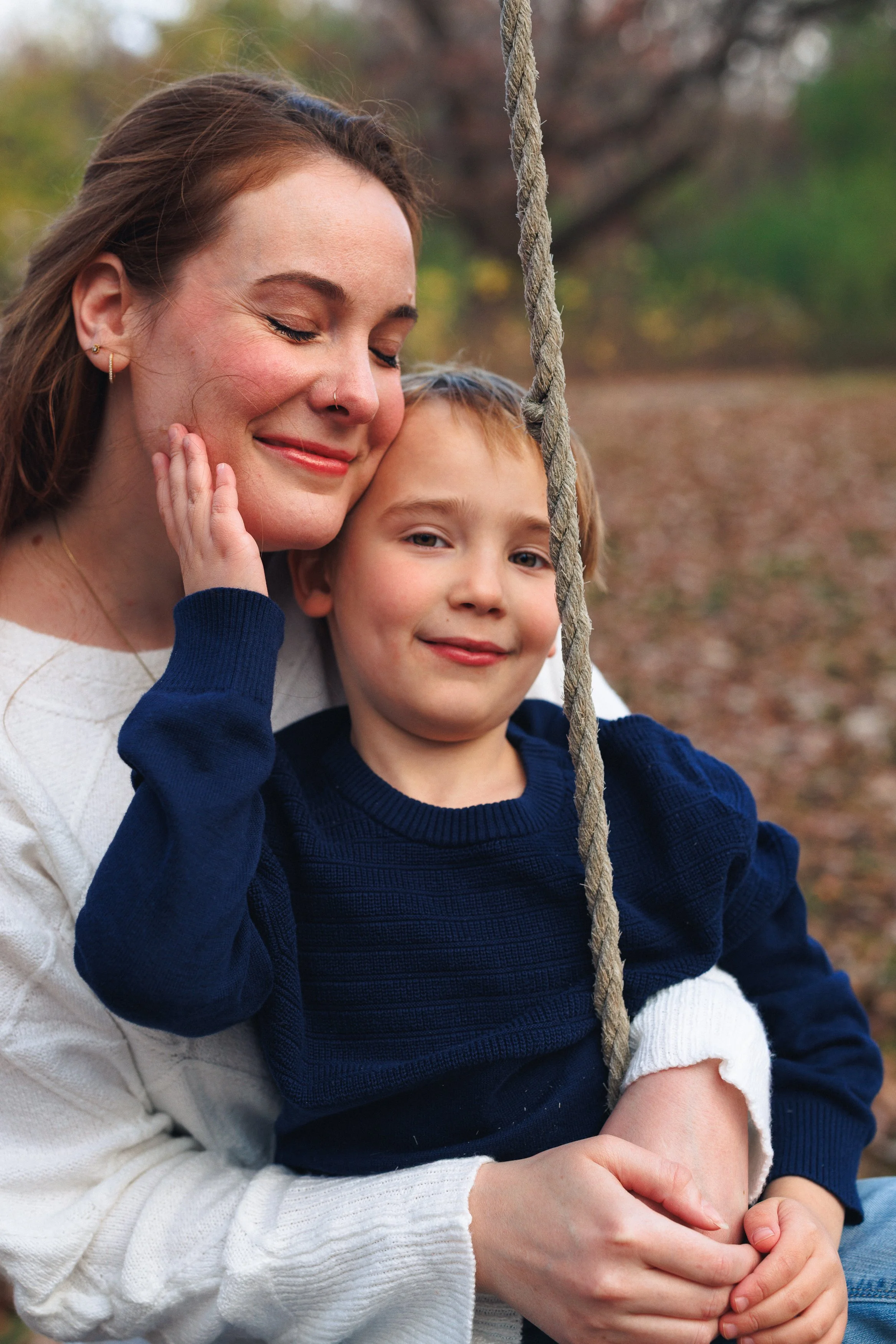 A smiling woman with closed eyes and a young boy sitting on a swing outdoors during autumn. The woman has light skin, reddish-brown hair, and is wearing a white sweater. The boy has light skin, light brown hair, and is wearing a navy blue sweater. A 